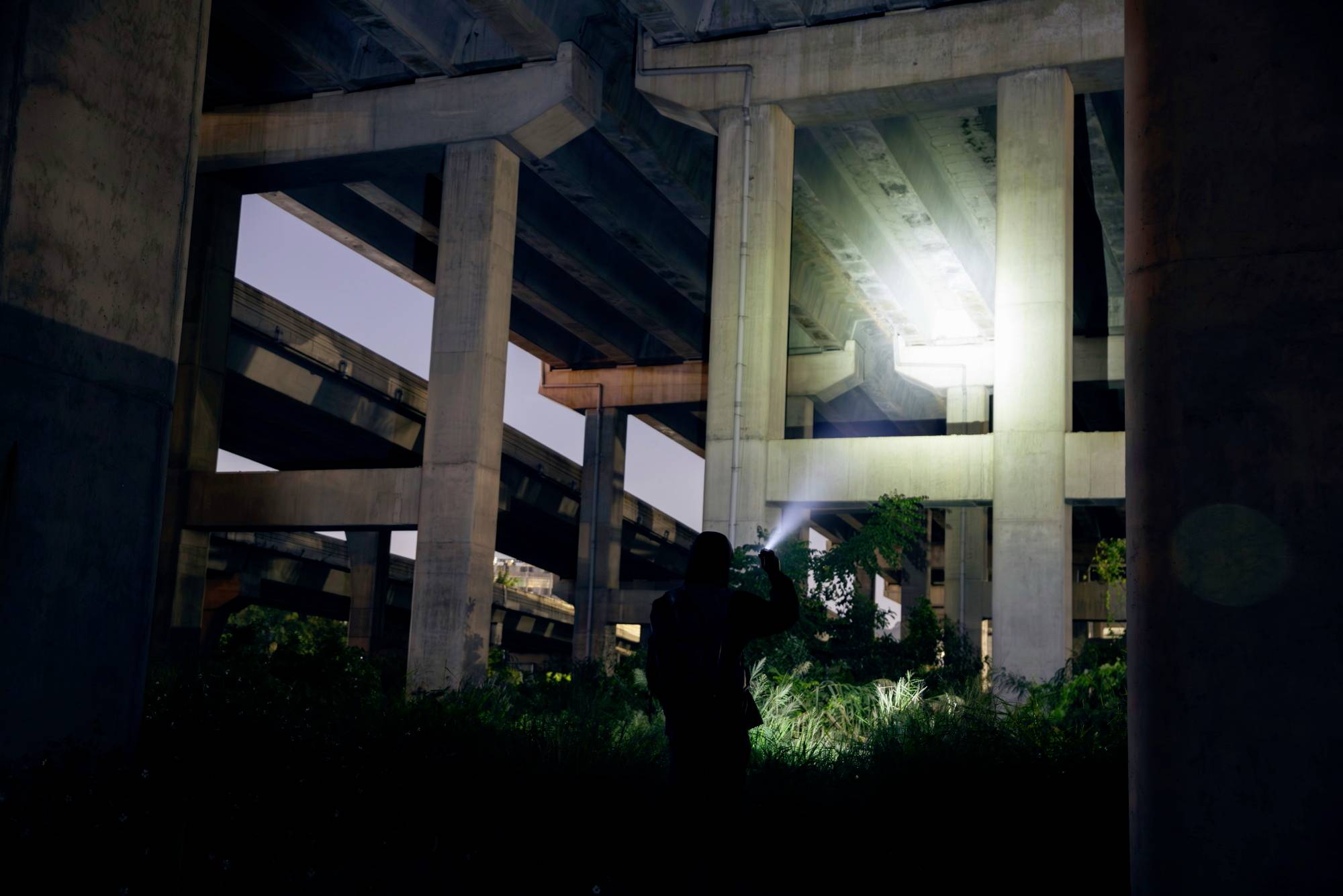 Une personne se tient la nuit sous un pont autoroutier et dirige la lumière d'une lampe de poche sur les piliers en béton.