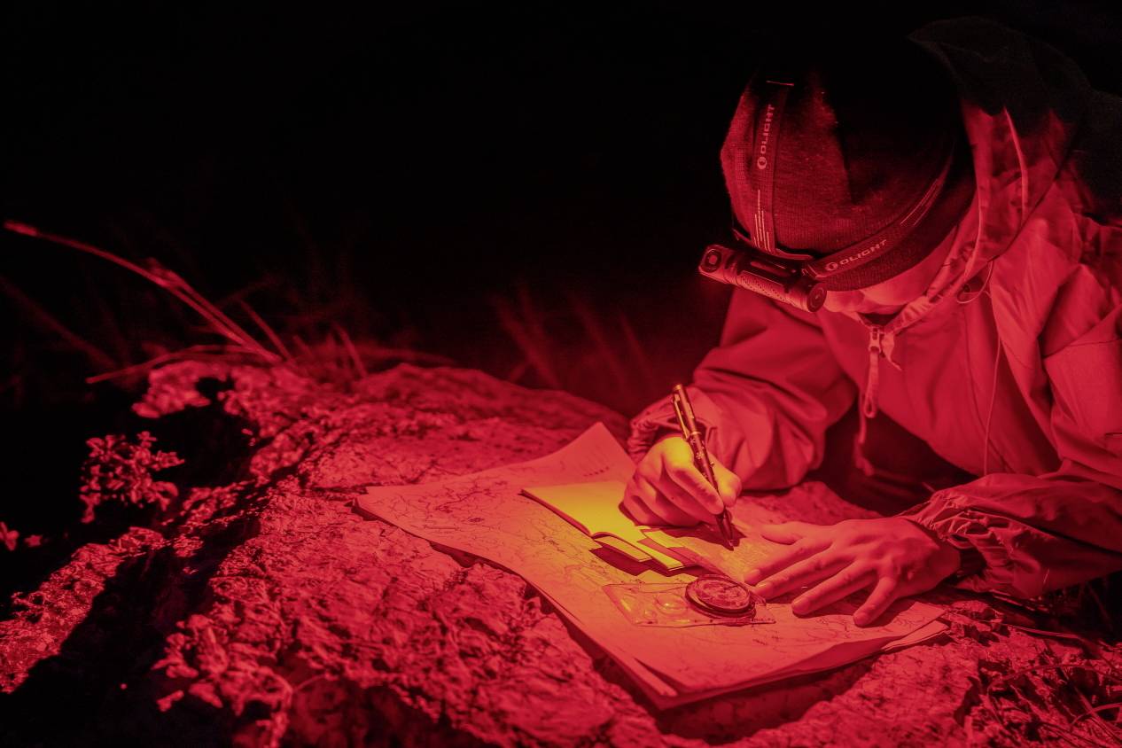 Une personne étudie une carte à la lumière rouge d'une lampe frontale dans l'obscurité. Elle prend des notes à côté d'elle avec une boussole.