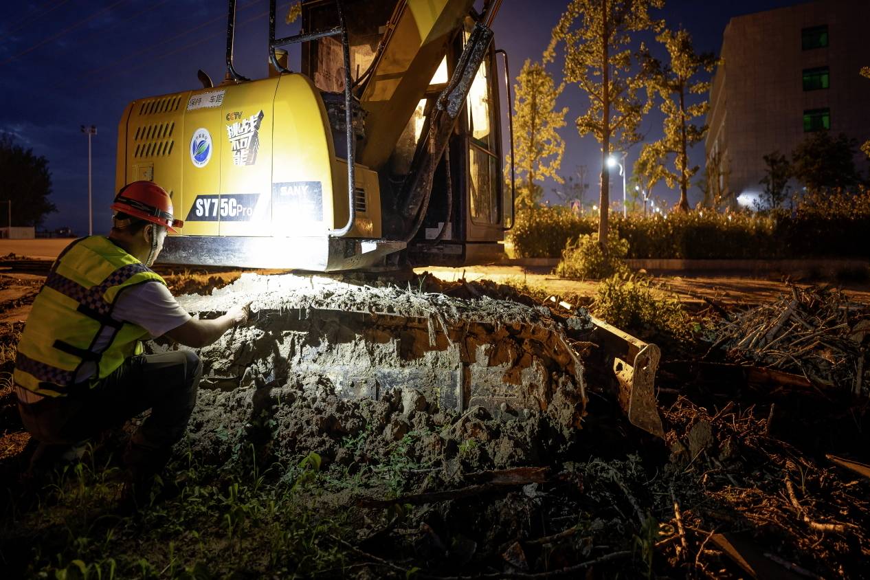 Une personne en tenue de travail vérifie la partie inférieure d'un engin de chantier jaune, éclairée par des projecteurs, sur un chantier de construction durant la nuit.