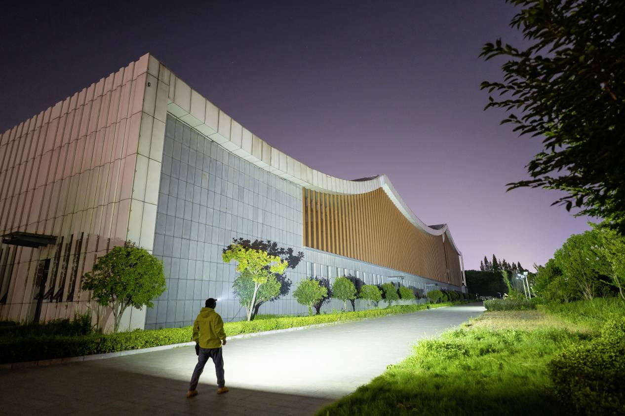 Une personne marche la nuit sur un trottoir éclairé longeant un grand bâtiment moderne. Des arbres bordent le chemin.