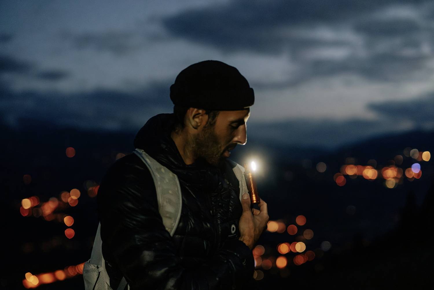 Une personne fixe du regard une lampe de poche dans l'obscurité, avec un paysage urbain illuminé par des lumières en arrière-plan.