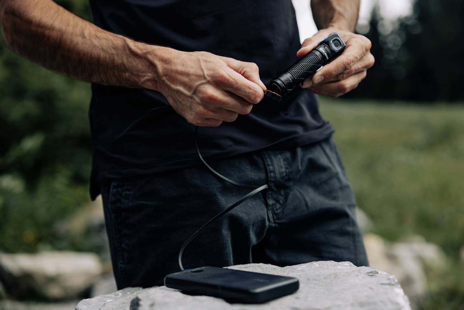 Une personne tient une lampe de poche et la relie à une batterie portable noire alors qu'elle se trouve à l'extérieur dans la nature.