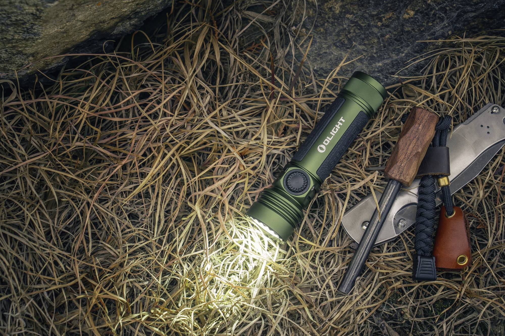 Une lampe de poche verte brille sur de l'herbe sèche à côté d'un couteau et d'un allume-feu sur un sol pierreux.