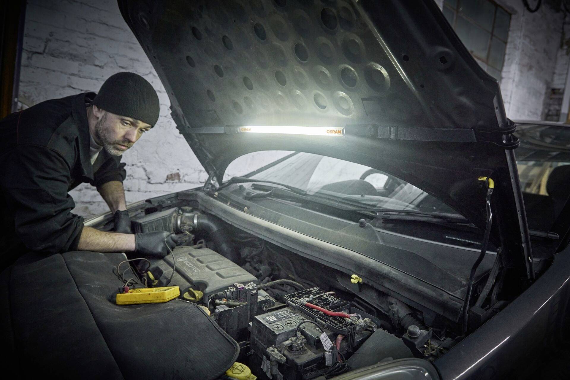 Un mécanicien vêtu de noir travaille avec un appareil de mesure sur le compartiment moteur ouvert d'une voiture dans un environnement d'atelier.