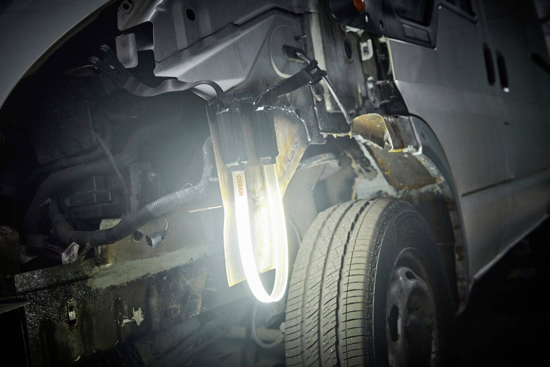 'Une voiture avec le capot ouvert révèle un châssis rouillé et un câblage apparent, éclairés par une lampe d'atelier.'
