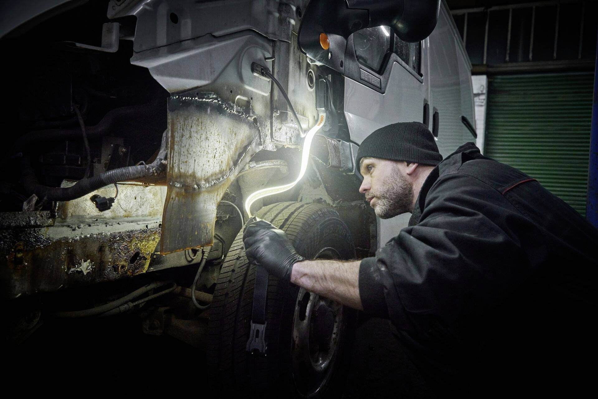 Un mécanicien en tenue de travail inspecte l'intérieur d'une voiture à l'aide d'une lampe d'inspection, concentré sur une pièce rouillée.