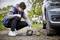 Un homme est agenouillé sur un chemin non revêtu et utilise un compresseur portable pour gonfler le pneu d'une voiture. Un vélo est visible en arrière-plan.