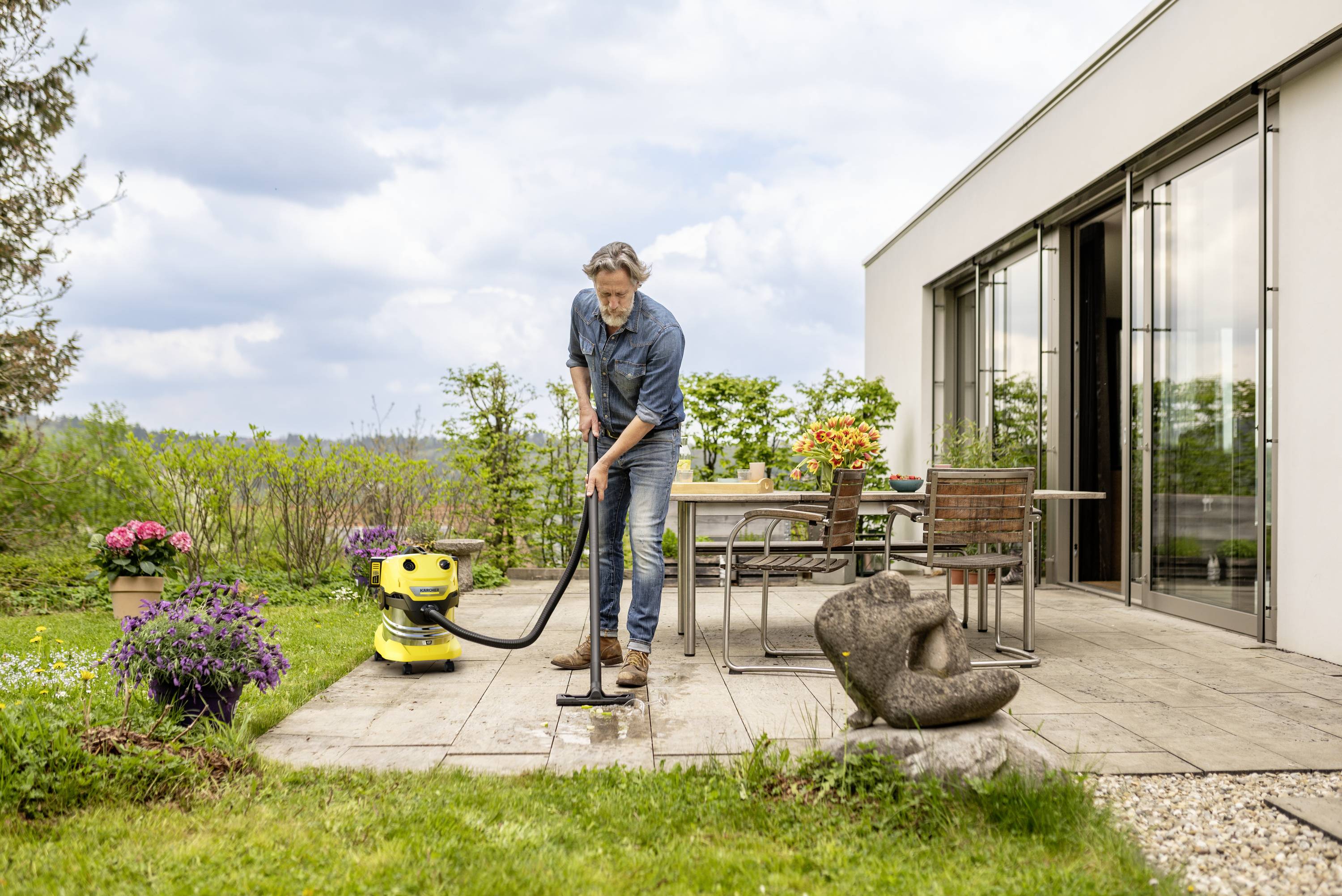 Un homme nettoie une terrasse avec un nettoyeur haute pression, entouré de plantes et de meubles de jardin. Le ciel est nuageux.