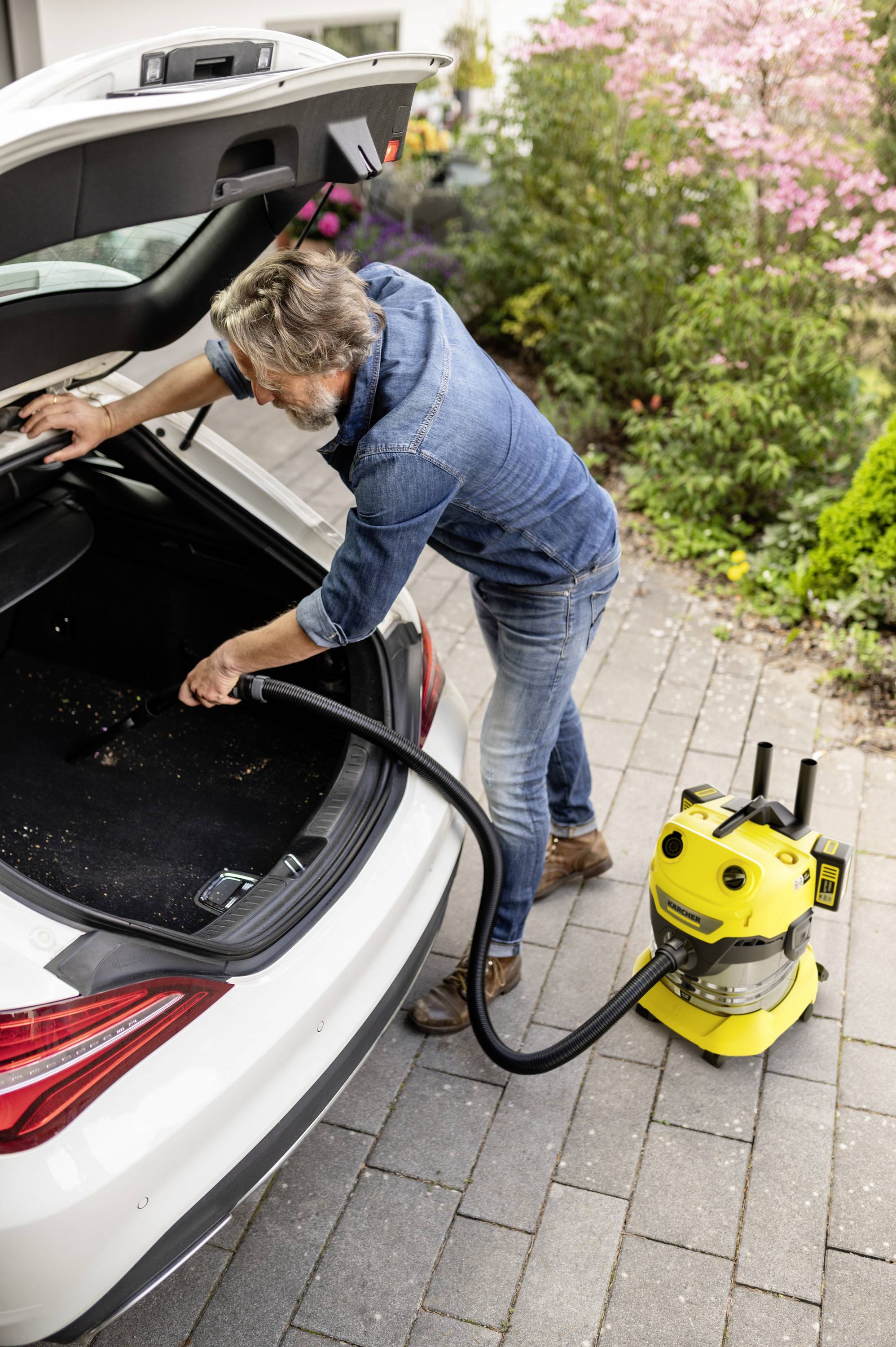 Un homme utilise un aspirateur jaune pour nettoyer le coffre d'une voiture blanche. Des fleurs sont visibles en arrière-plan.
