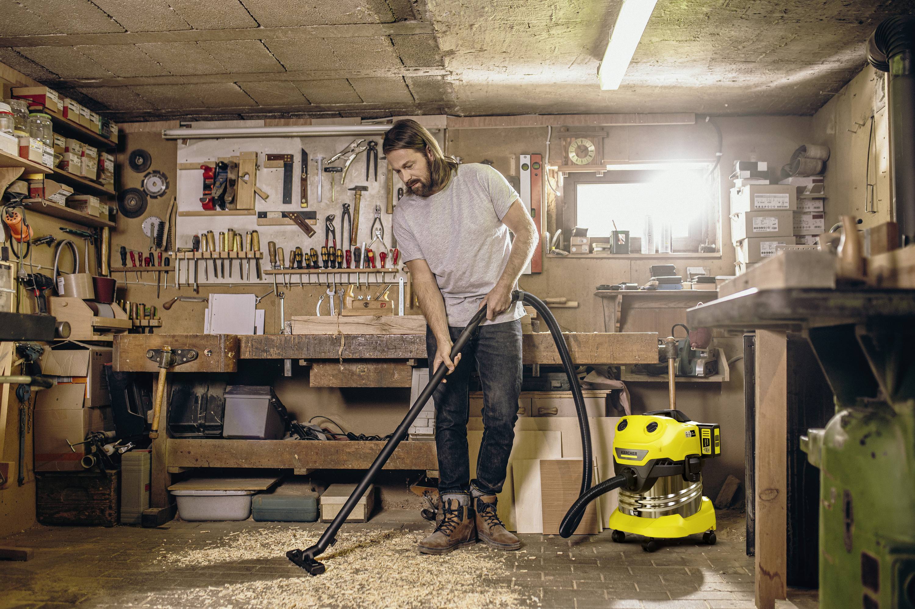 Un homme aspire des copeaux de bois dans un atelier. Des outils sont accrochés au mur, et un aspirateur jaune est posé à côté de lui.