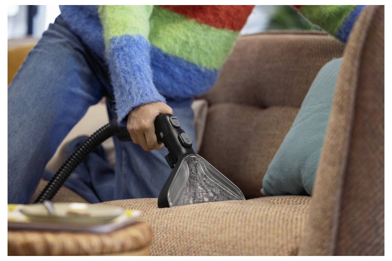 Une personne portant un pull coloré utilise un aspirateur à main sur un canapé brun, illustrant le nettoyage domestique.