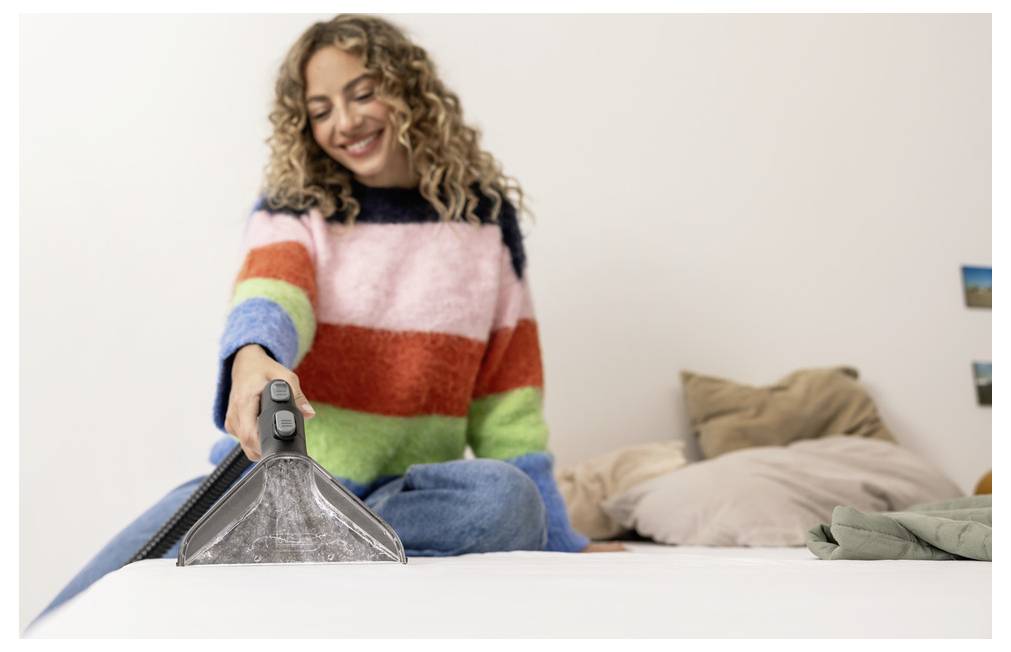 Une personne portant un pull coloré utilise un aspirateur sur un lit blanc, en souriant. Des oreillers sont visibles en arrière-plan.