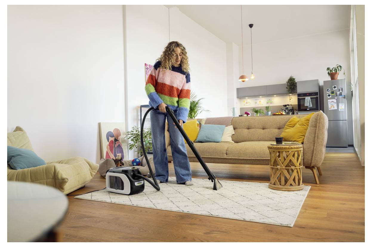 Une personne passe l'aspirateur dans un salon chaleureux au design moderne, avec un canapé beige, des coussins colorés, un tapis à motifs et une lampe élégante.