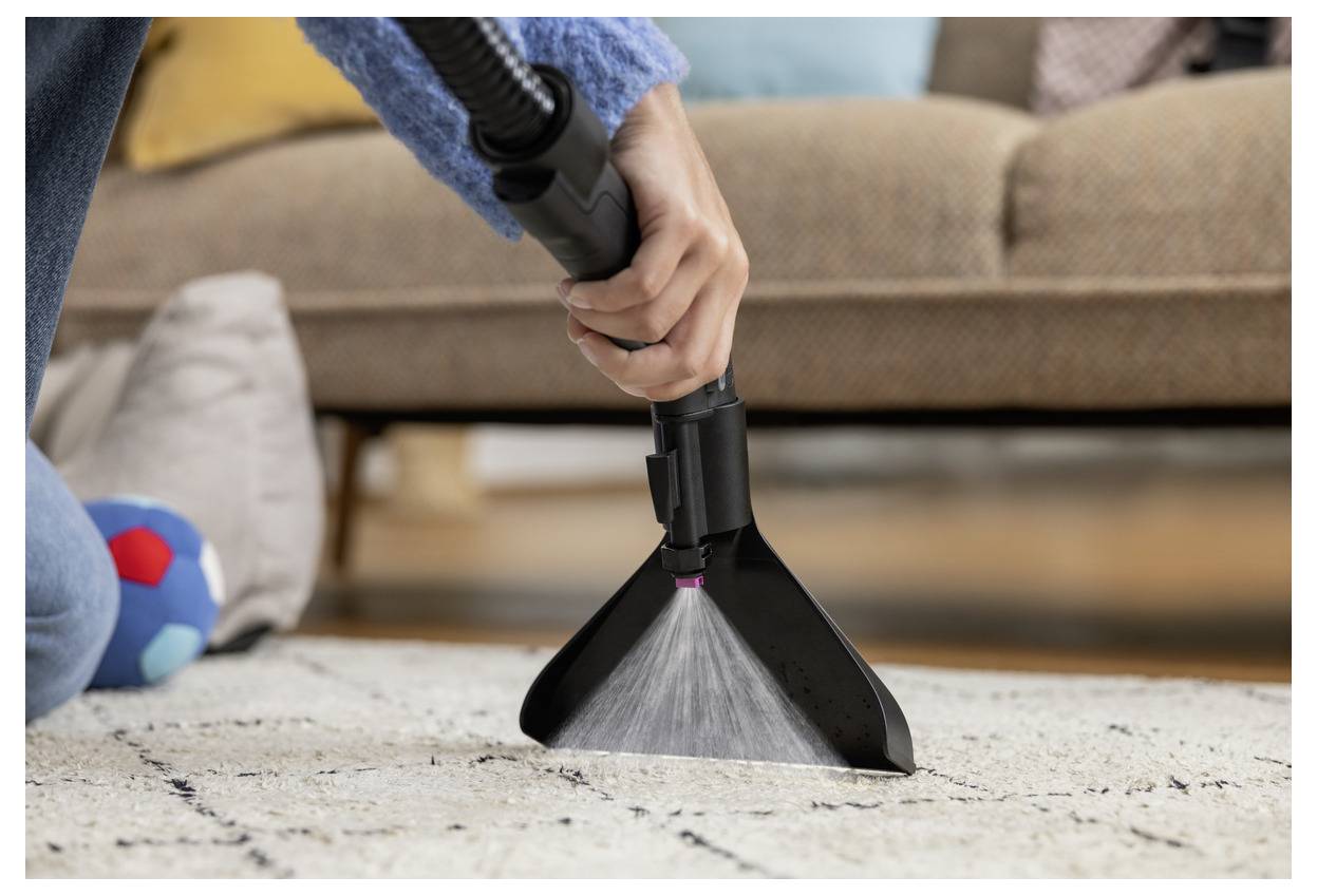 Une personne aspire un tapis avec un accessoire d'aspirateur à main, en se concentrant sur l'élimination des saletés près d'un canapé et d'un ballon coloré.