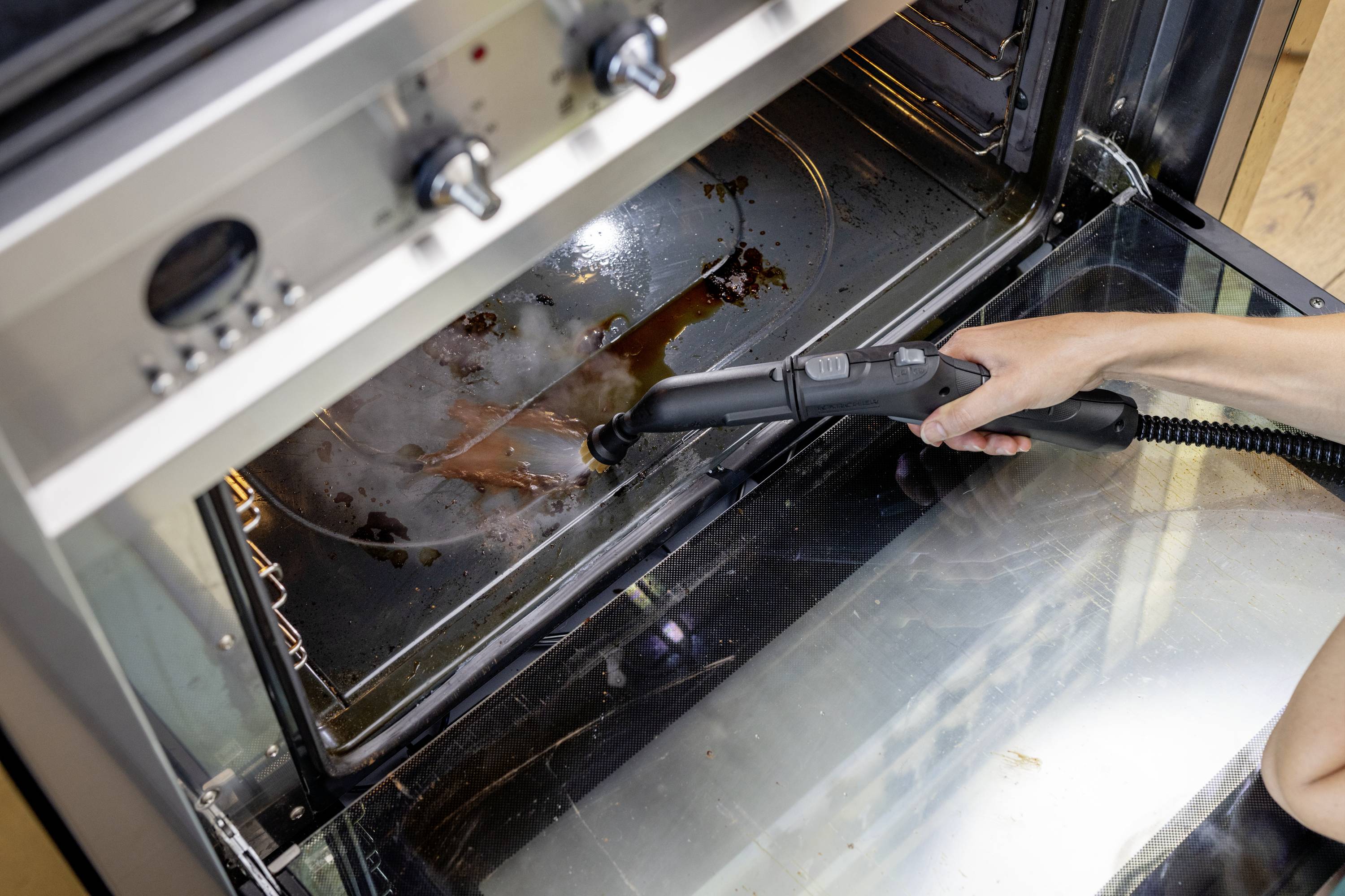 Une personne nettoie un four sale à l'aide d'un nettoyeur vapeur. Des restes de nourriture brûlés sur le fond du four sont en train d'être éliminés.