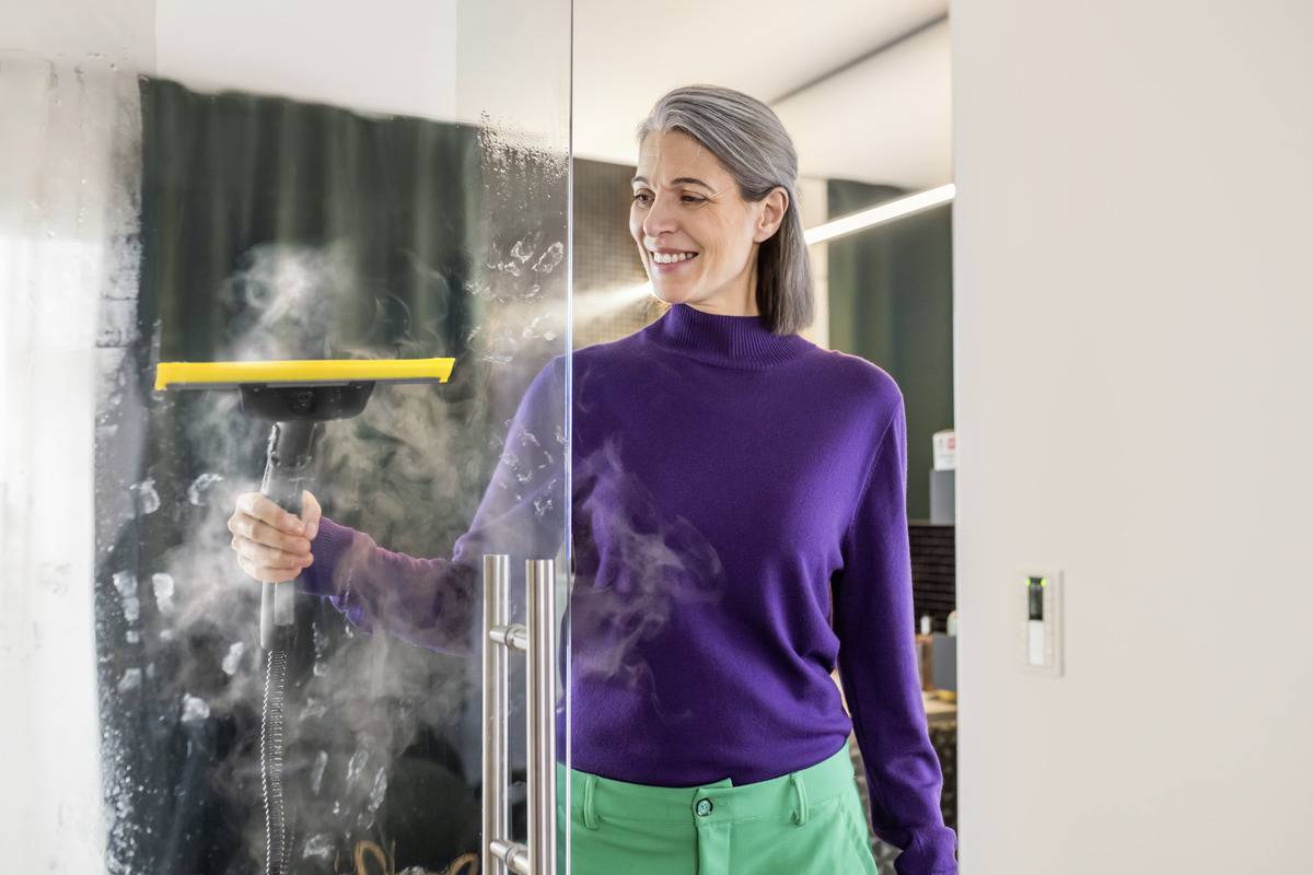 Une femme aux cheveux gris, portant un pull violet, nettoie une porte de douche en verre à l'aide d'un nettoyeur vapeur. De la vapeur s'élève tandis qu'elle travaille.