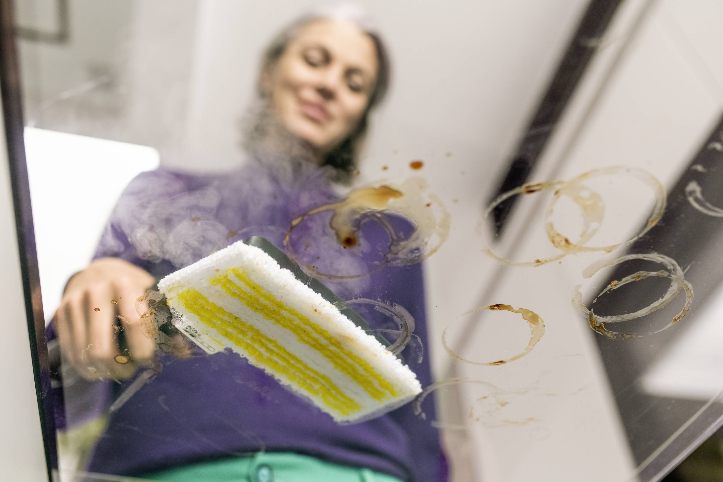 Une personne nettoie une surface vitrée sale à l'aide d'une éponge, vue depuis en dessous de la vitre.