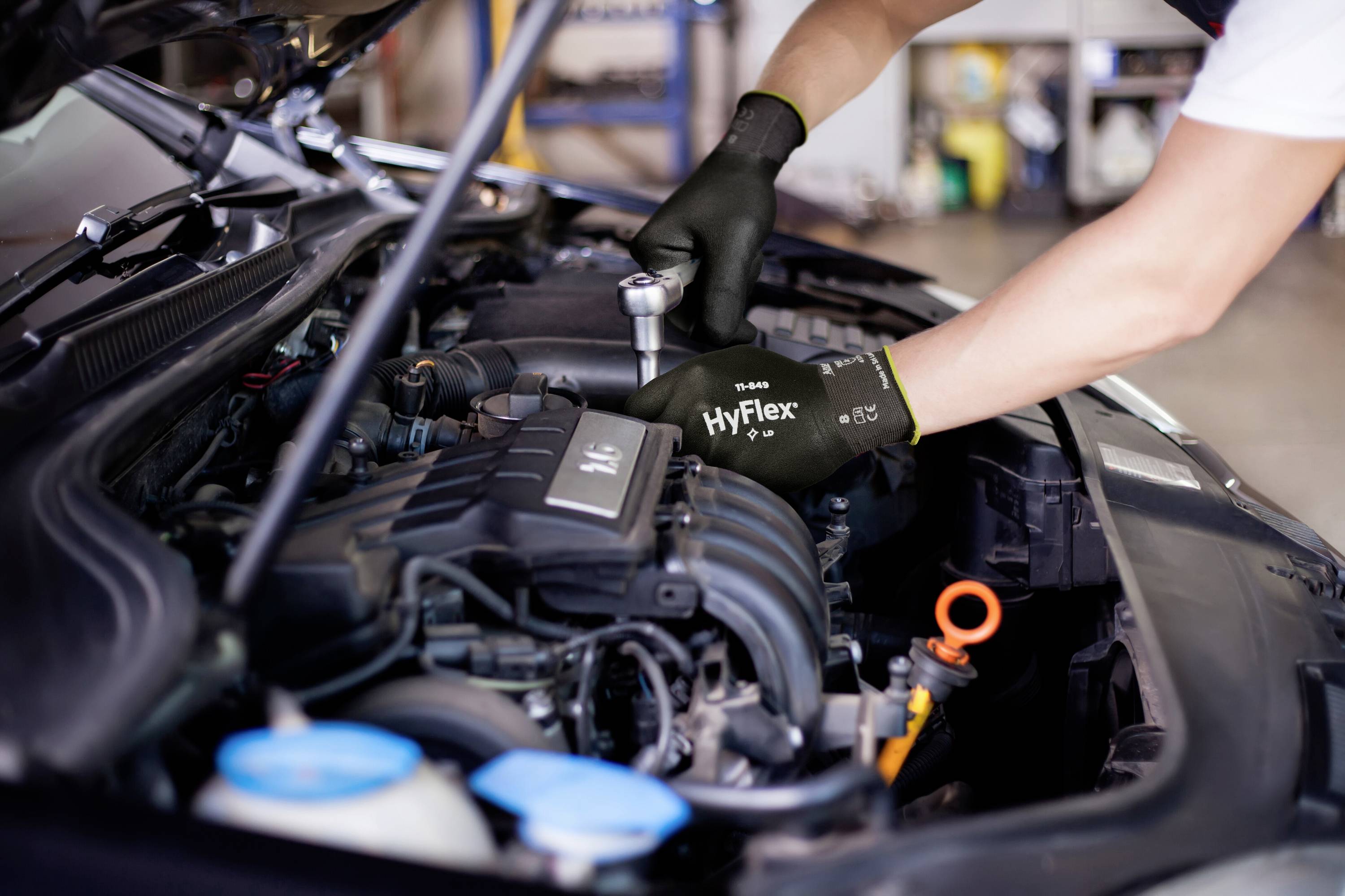 Une personne portant des gants travaille sur le moteur d'une voiture dont le capot est ouvert. Des outils et des liquides moteur sont visibles au premier plan.