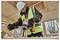 Un ouvrier du bâtiment portant un gilet de sécurité haute visibilité et un casque utilise une perceuse électrique sur une surface en bois à l'intérieur d'un bâtiment en construction.