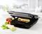 Un grill à contact ouvert avec un panini et des légumes frais à côté sur un plan de travail de cuisine.