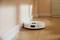 Un robot aspirateur blanc nettoie le sol en bois le long d'un mur dans une pièce lumineuse.