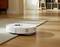 Un robot aspirateur blanc passe d'un parquet en bois à un tapis. L'arrière-plan montre un intérieur de salon chaleureux.