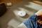Un robot aspirateur blanc nettoie des miettes sur un tapis dans un salon avec des canapés et des couvertures en arrière-plan.