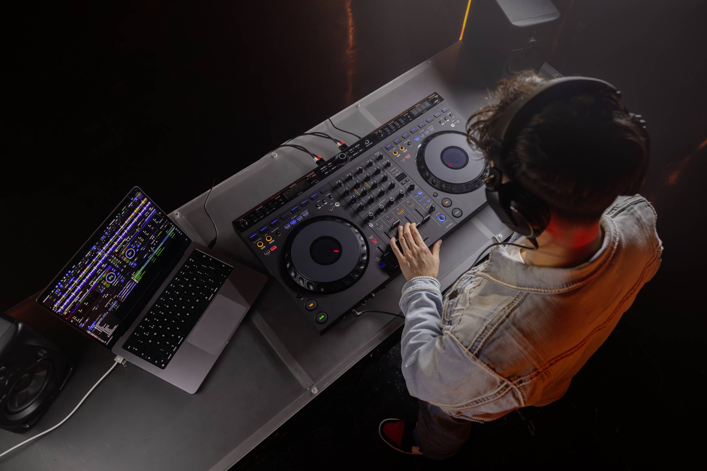 Un DJ mixe de la musique sur une table de mixage, un ordinateur portable ouvert à côté avec un logiciel de musique. Il porte des écouteurs et est concentré sur son travail.