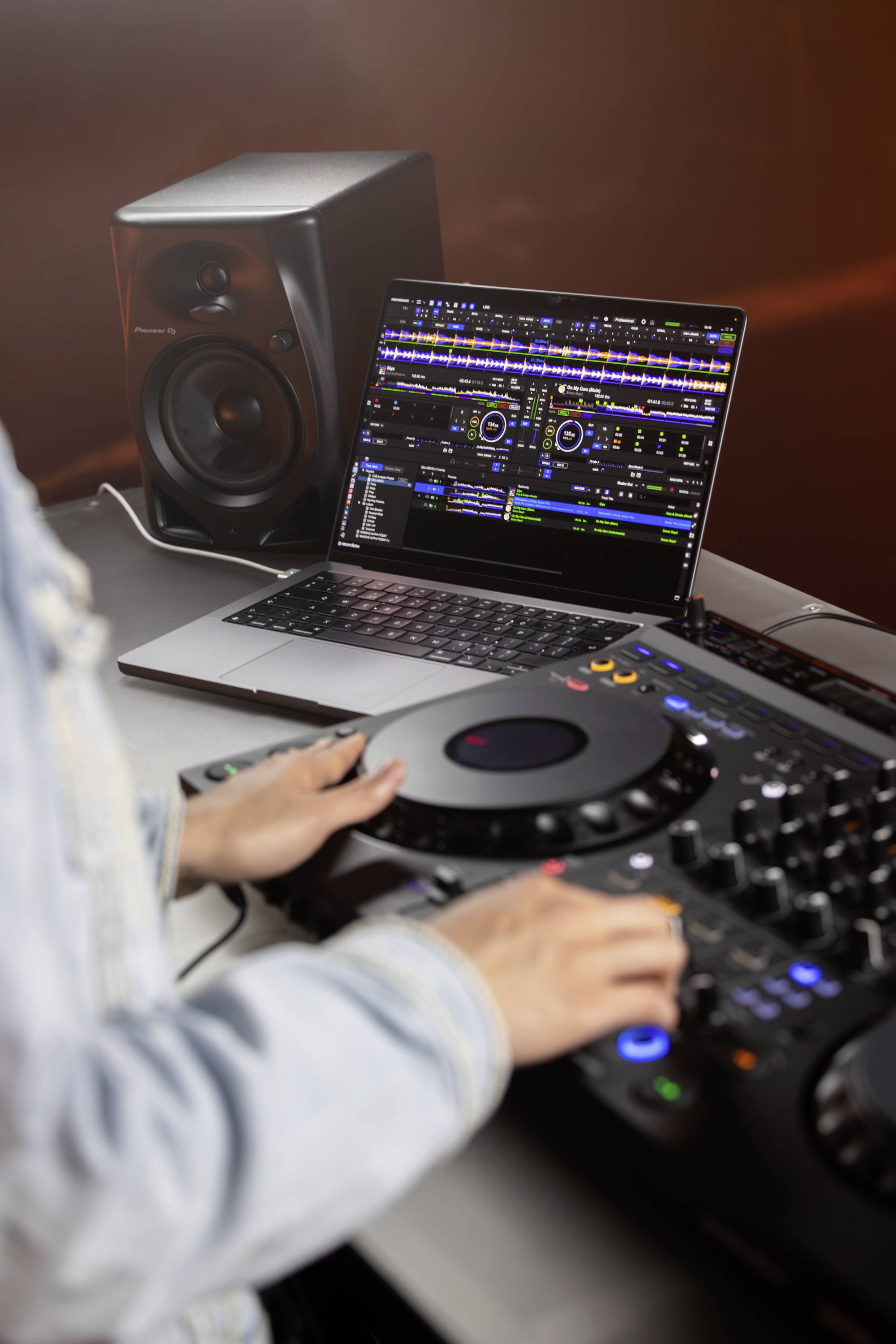 Une personne manipule une table de mixage de DJ. Sur l'écran de l'ordinateur portable, on peut voir des applications de logiciels musicaux. Un haut-parleur est placé en arrière-plan.