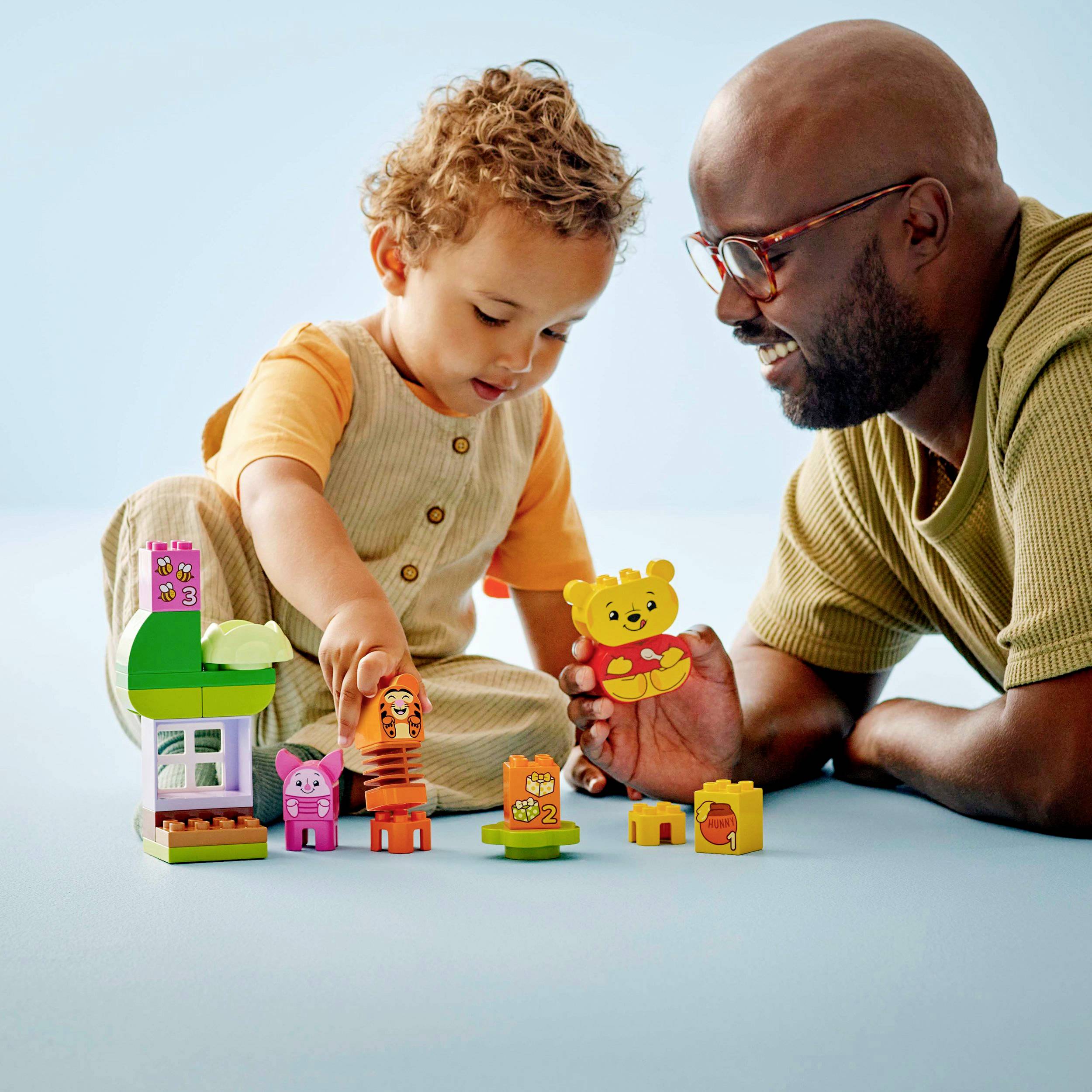 Un jeune enfant riant et un adulte jouent ensemble avec des blocs de construction colorés en forme de porte et des figurines sur une table bleue.