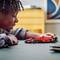 Un enfant sourit en jouant avec une voiture de course rouge jouet sur une table. Des meubles et des décorations flous sont visibles en arrière-plan.