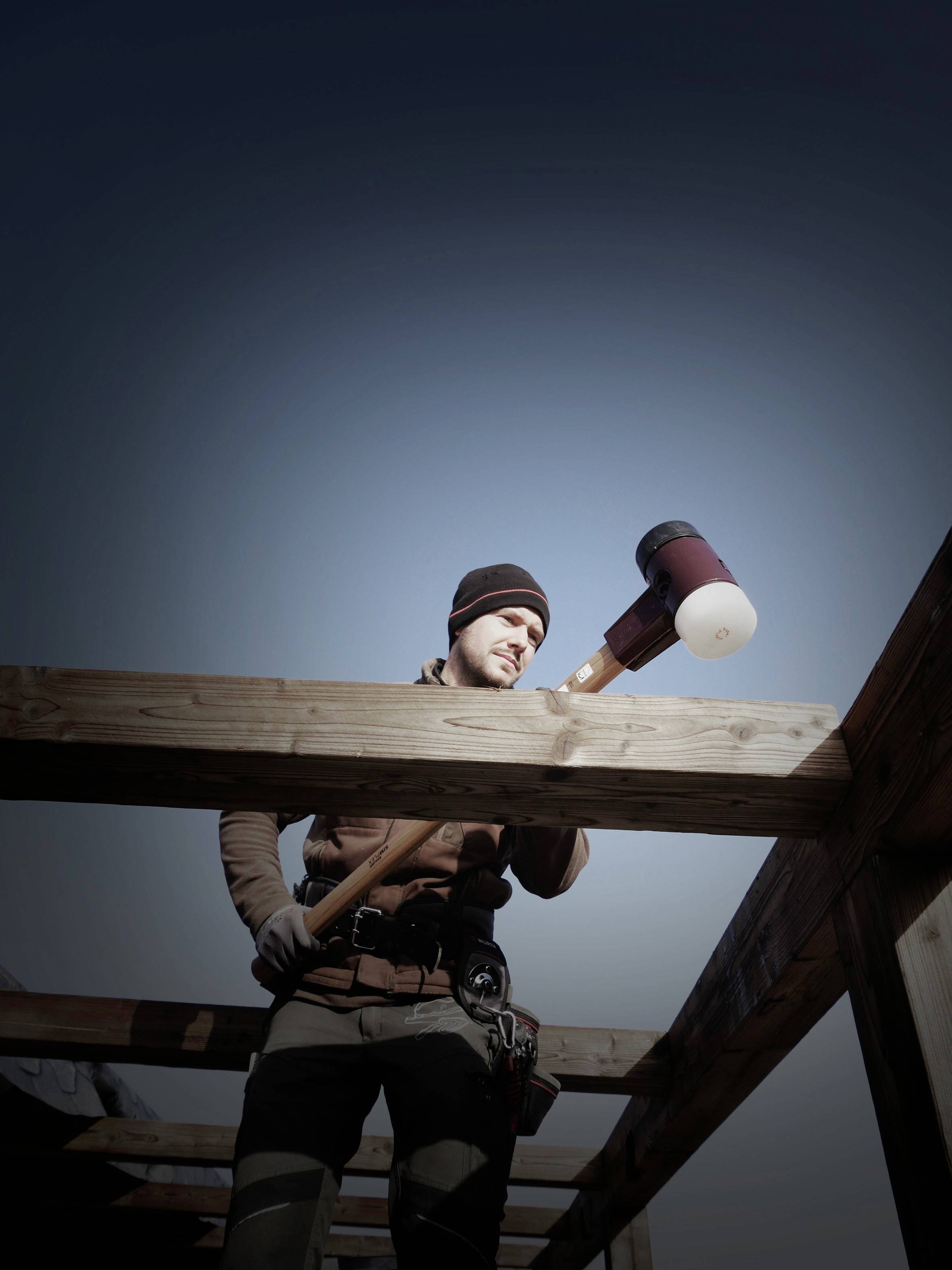 Un ouvrier portant une casquette et des gants utilise un grand marteau pour travailler une structure en bois à l'extérieur. Le ciel est dégagé.