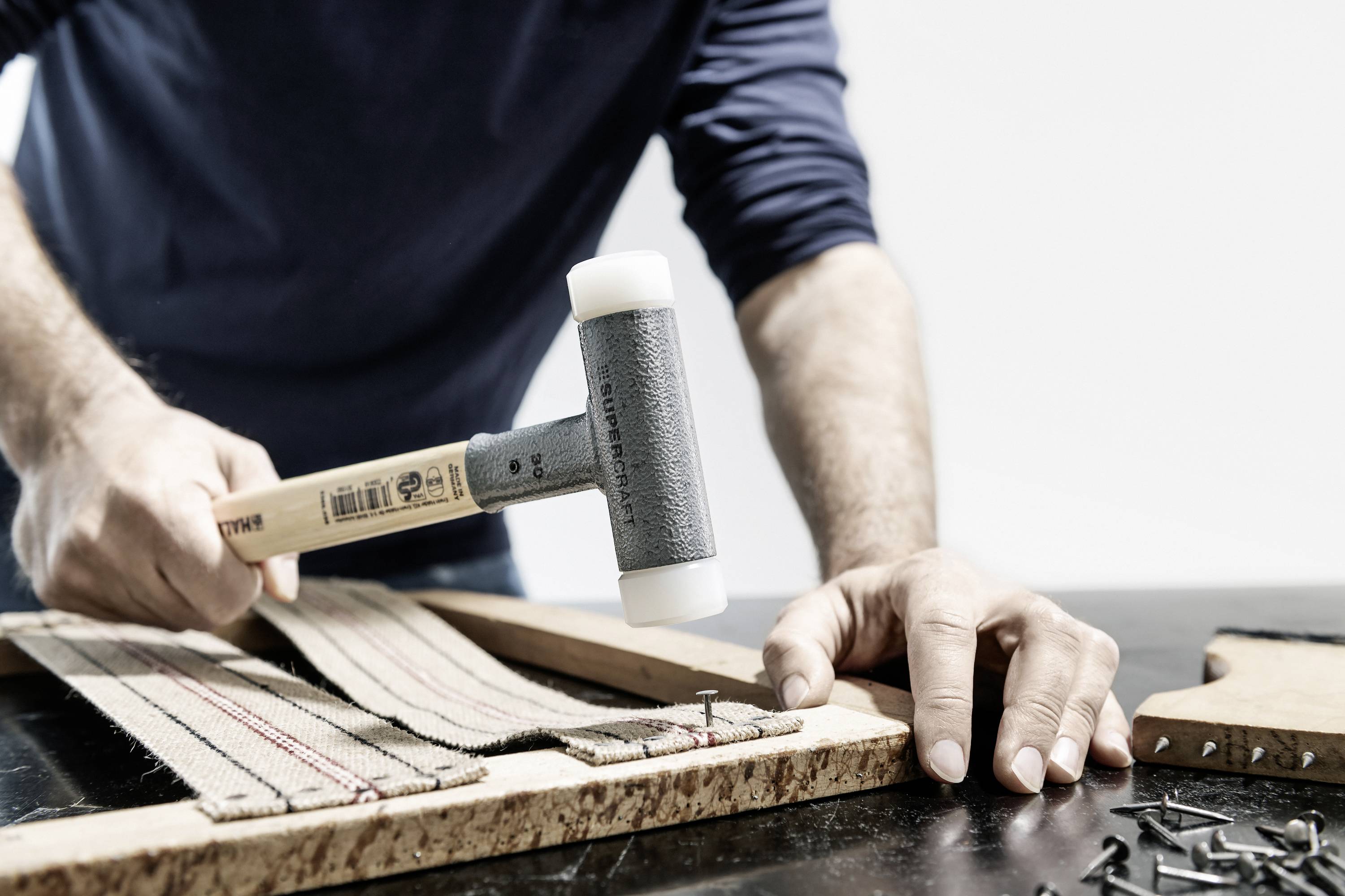 Une personne frappe avec un marteau en plastique sur un tissu fixé à un morceau de bois rectangulaire. Des clous sont prêts à être utilisés.