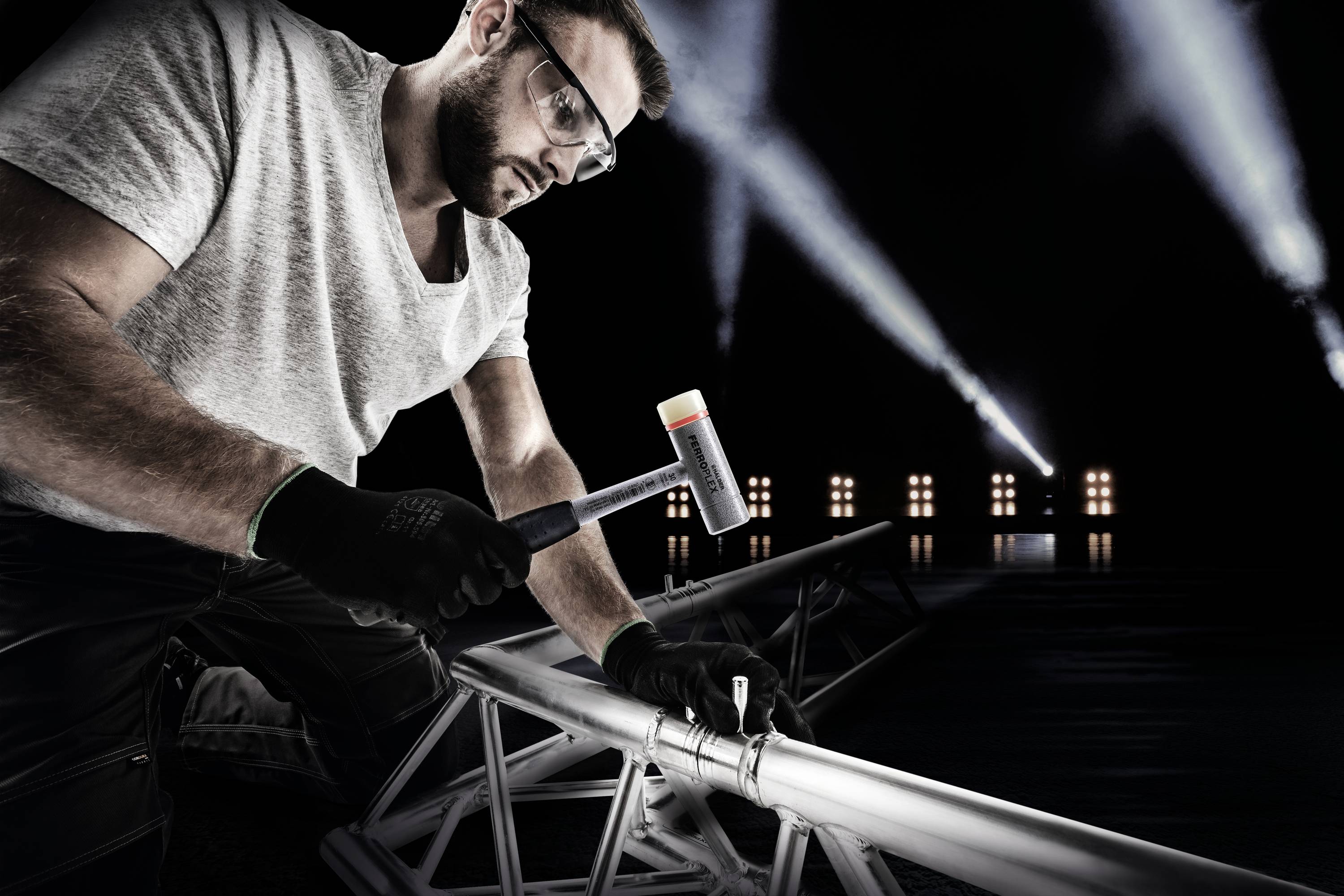 Un homme portant des lunettes et des gants frappe un échafaudage métallique avec un marteau, tandis que des projecteurs lumineux brillent en arrière-plan.