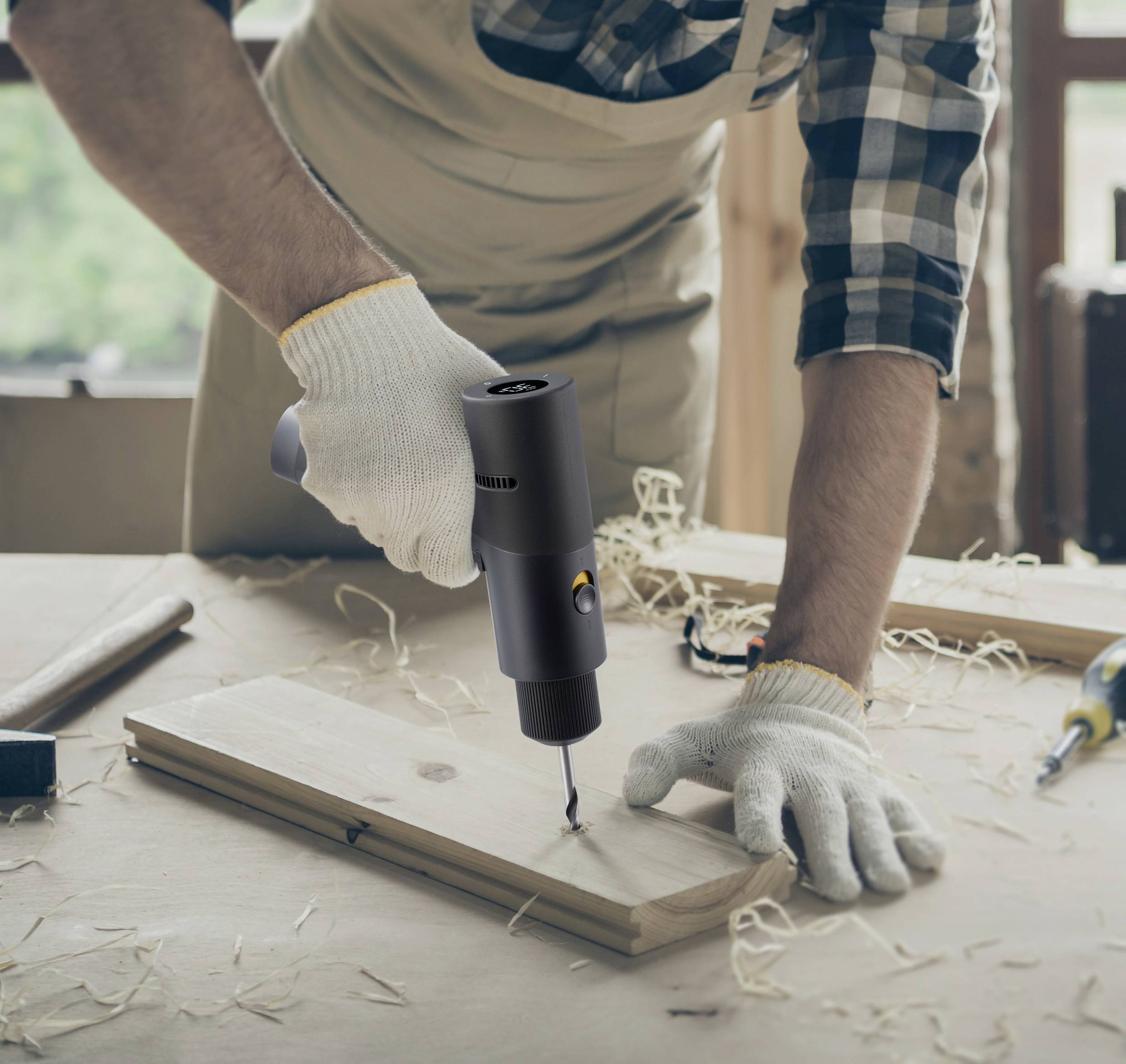 Une personne fore un trou dans une planche en bois à l'aide d'une perceuse électrique dans un environnement d'atelier. Des gants protègent les mains.