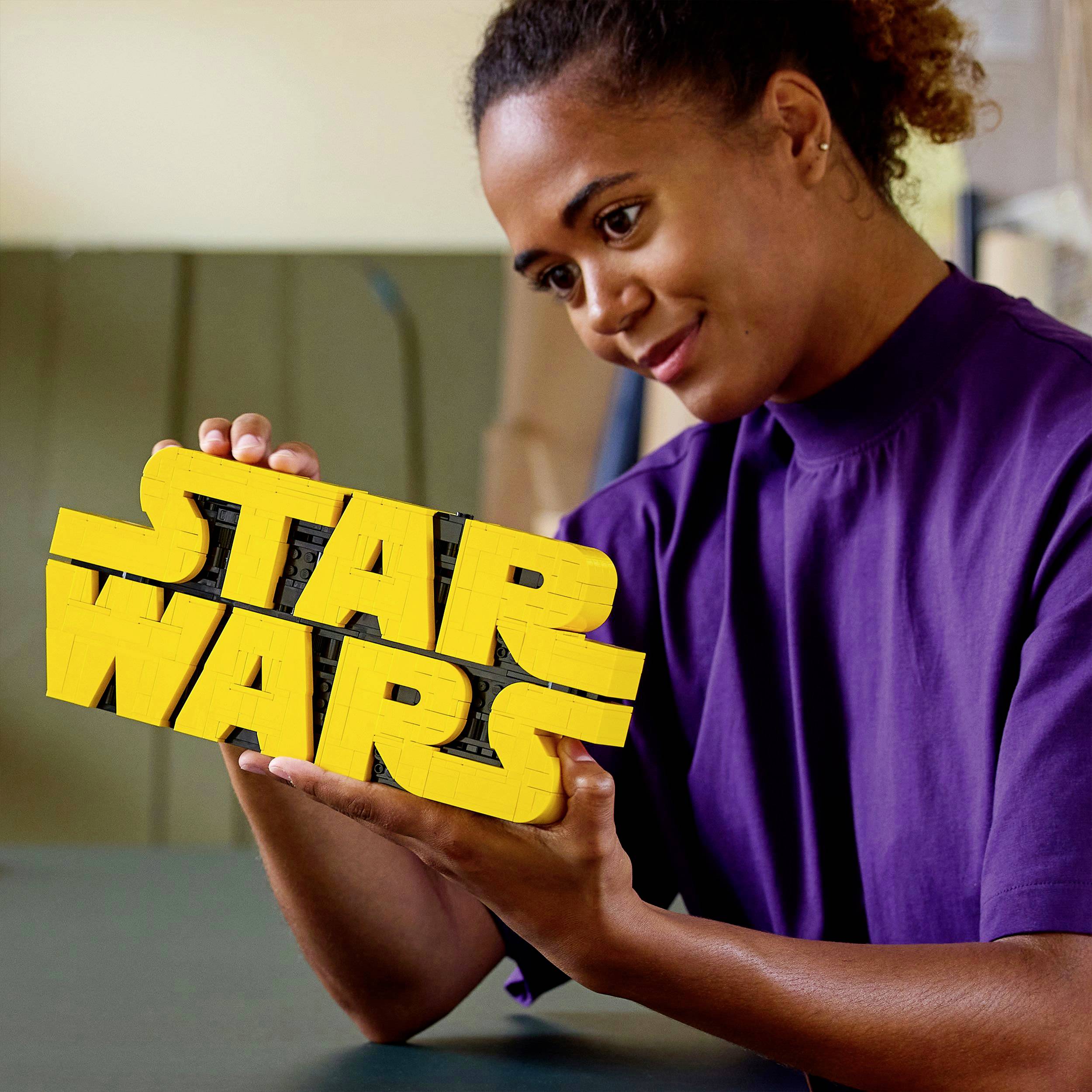 Une personne sourit en tenant un modèle jaune 'STAR WARS' dans ses mains, assise à une table.