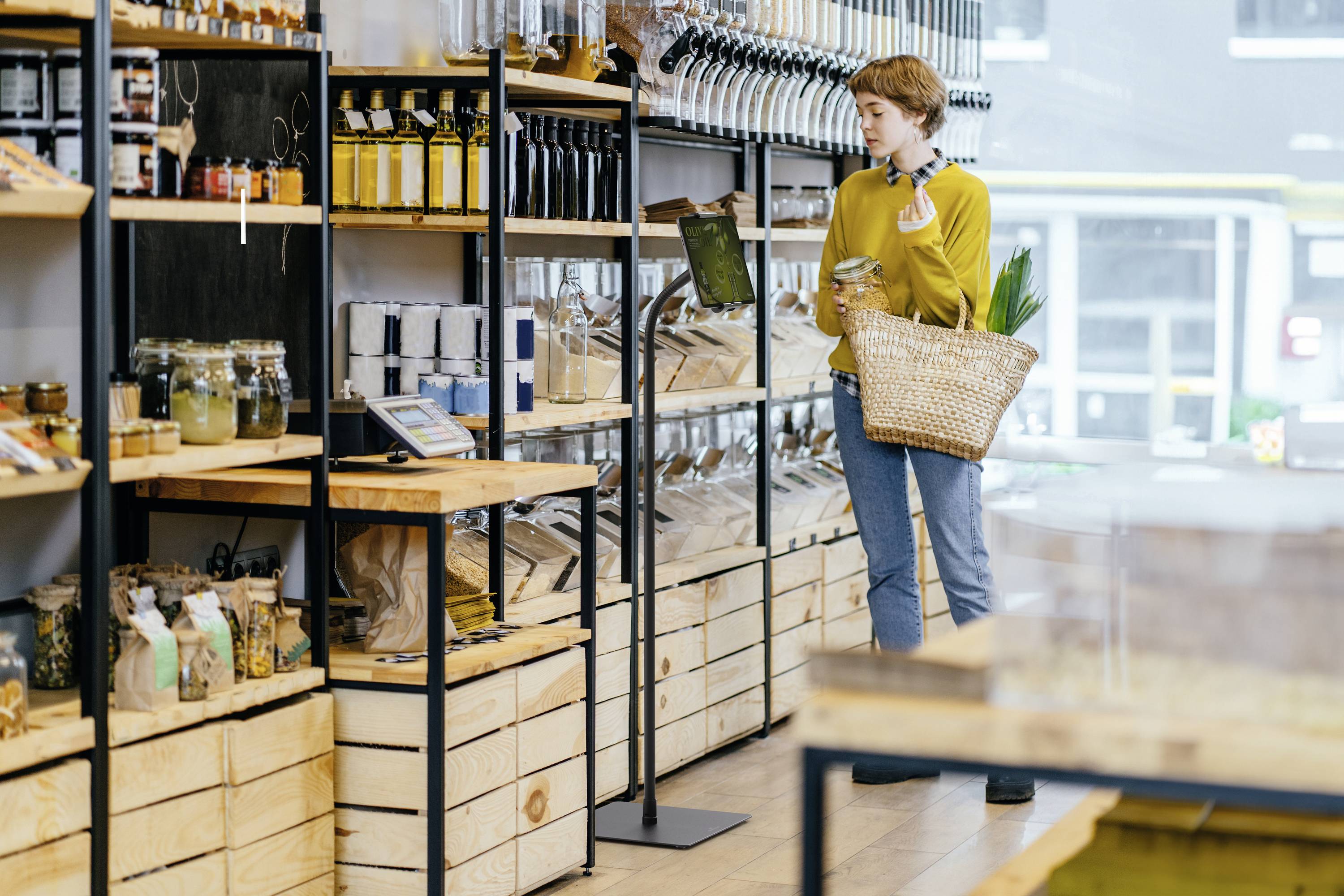 Une personne se tient dans une boutique moderne avec des paniers et des étagères remplis de bocaux et de sacs. Elle examine attentivement un produit.