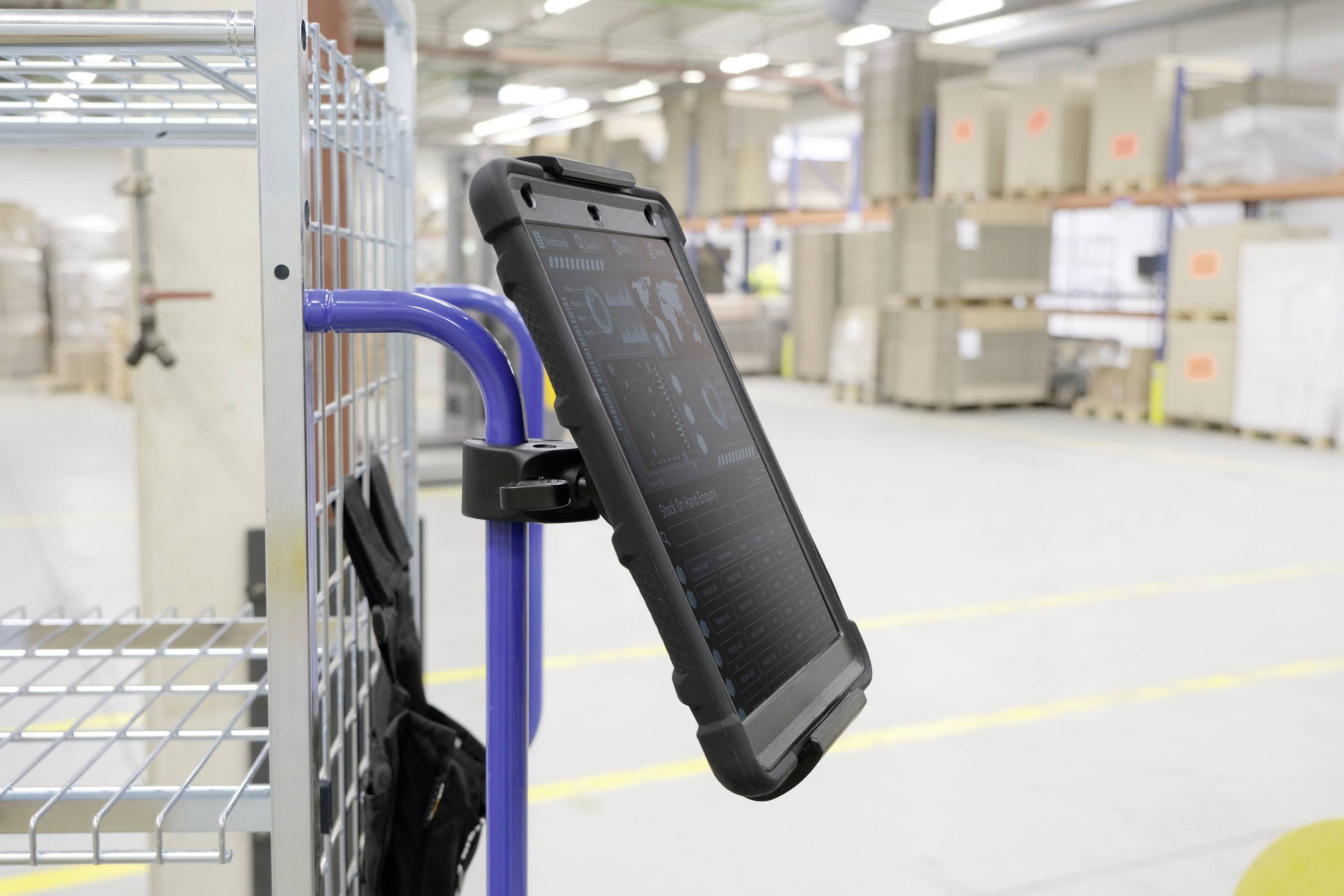 Une tablette fixée à un chariot de manutention dans un entrepôt, affichant des informations techniques sur son écran. Des étagères et des cartons sont visibles en arrière-plan.