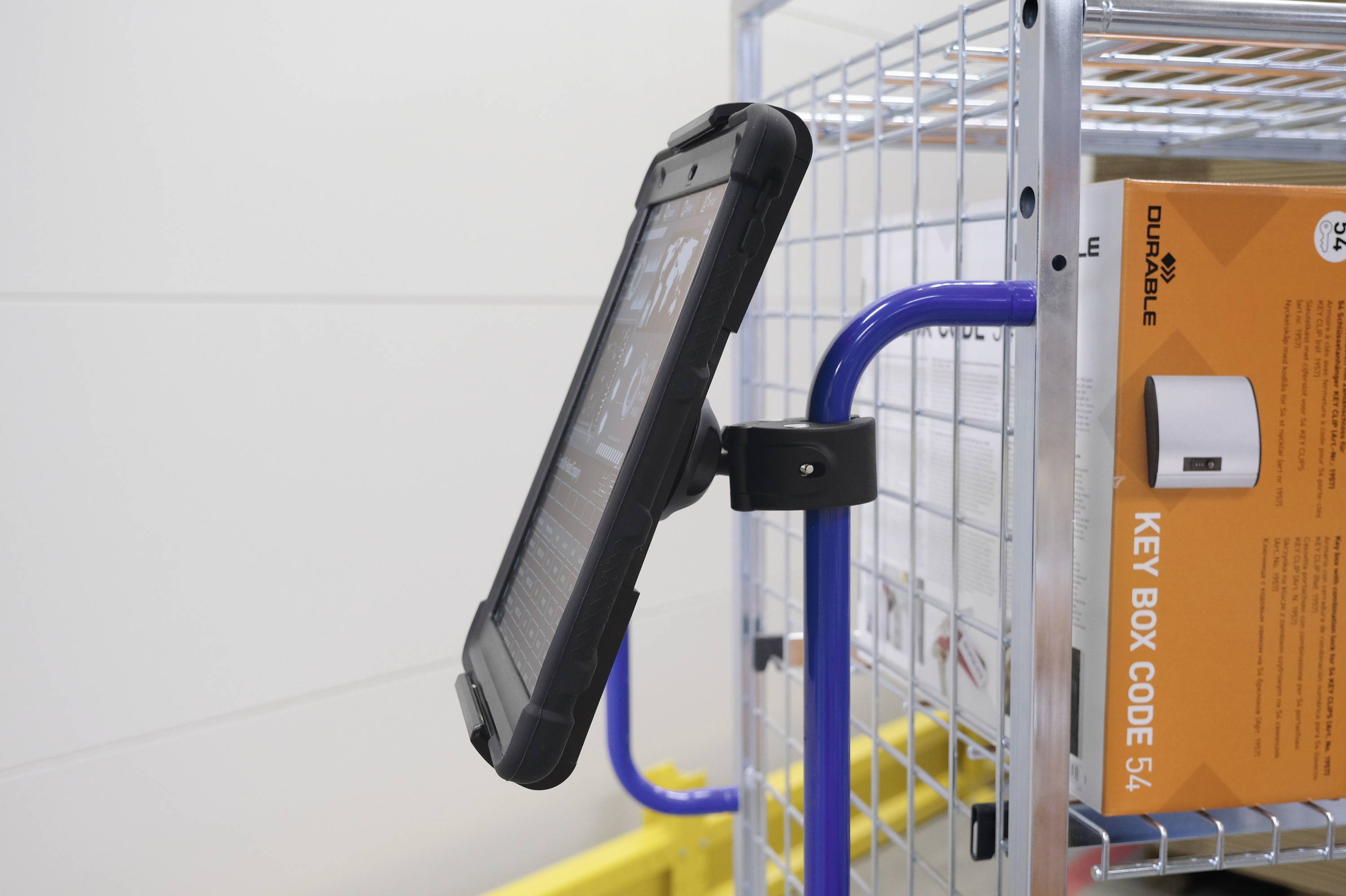 Une tablette est fixée dans un support sur un chariot métallique. À côté se trouve un carton orange portant l'inscription 'KEY BOX CODE 54'.