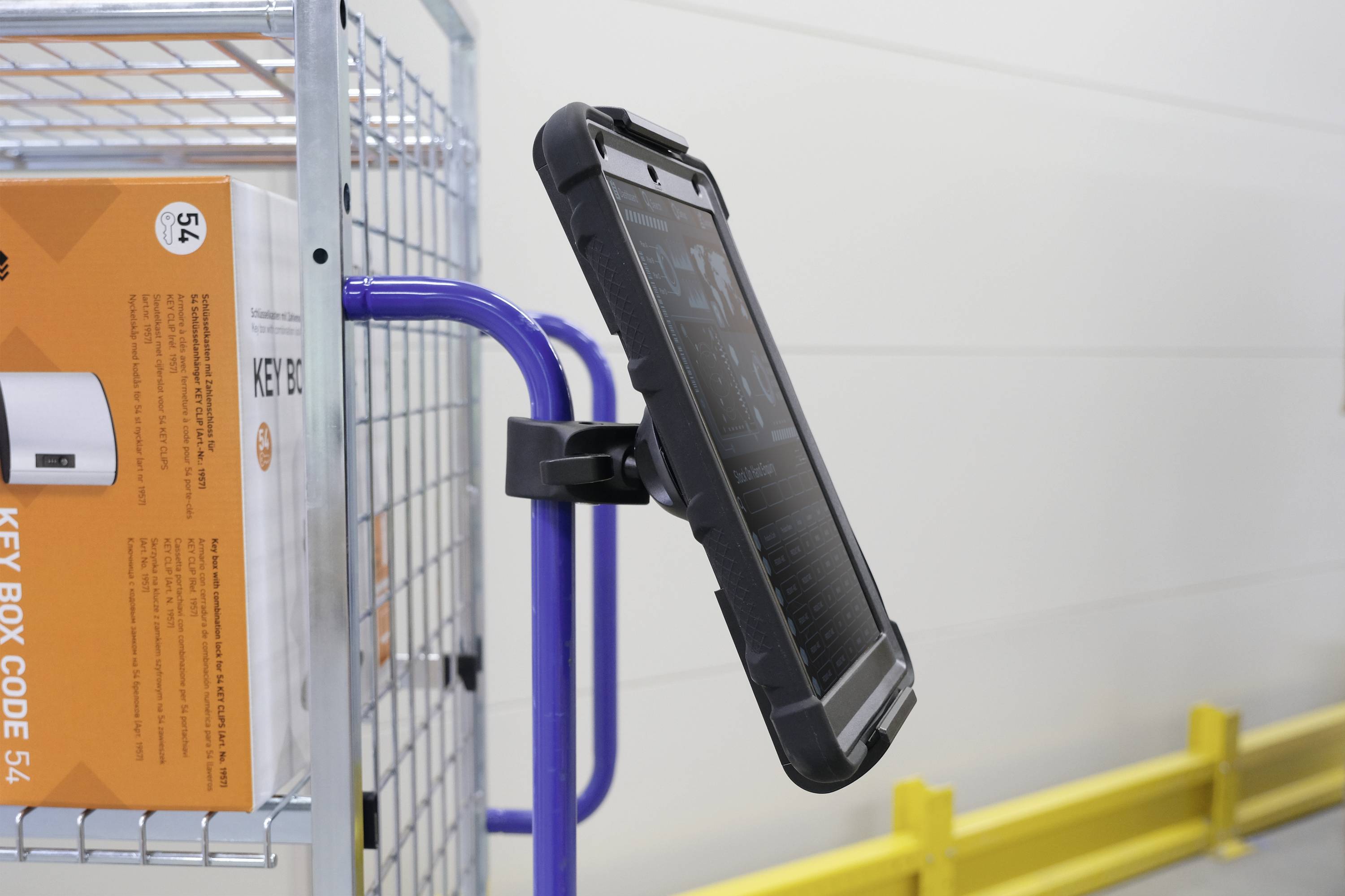 Une tablette dans un étui robuste est fixée sur un support métallique bleu à côté d'une étagère avec des boîtes oranges.