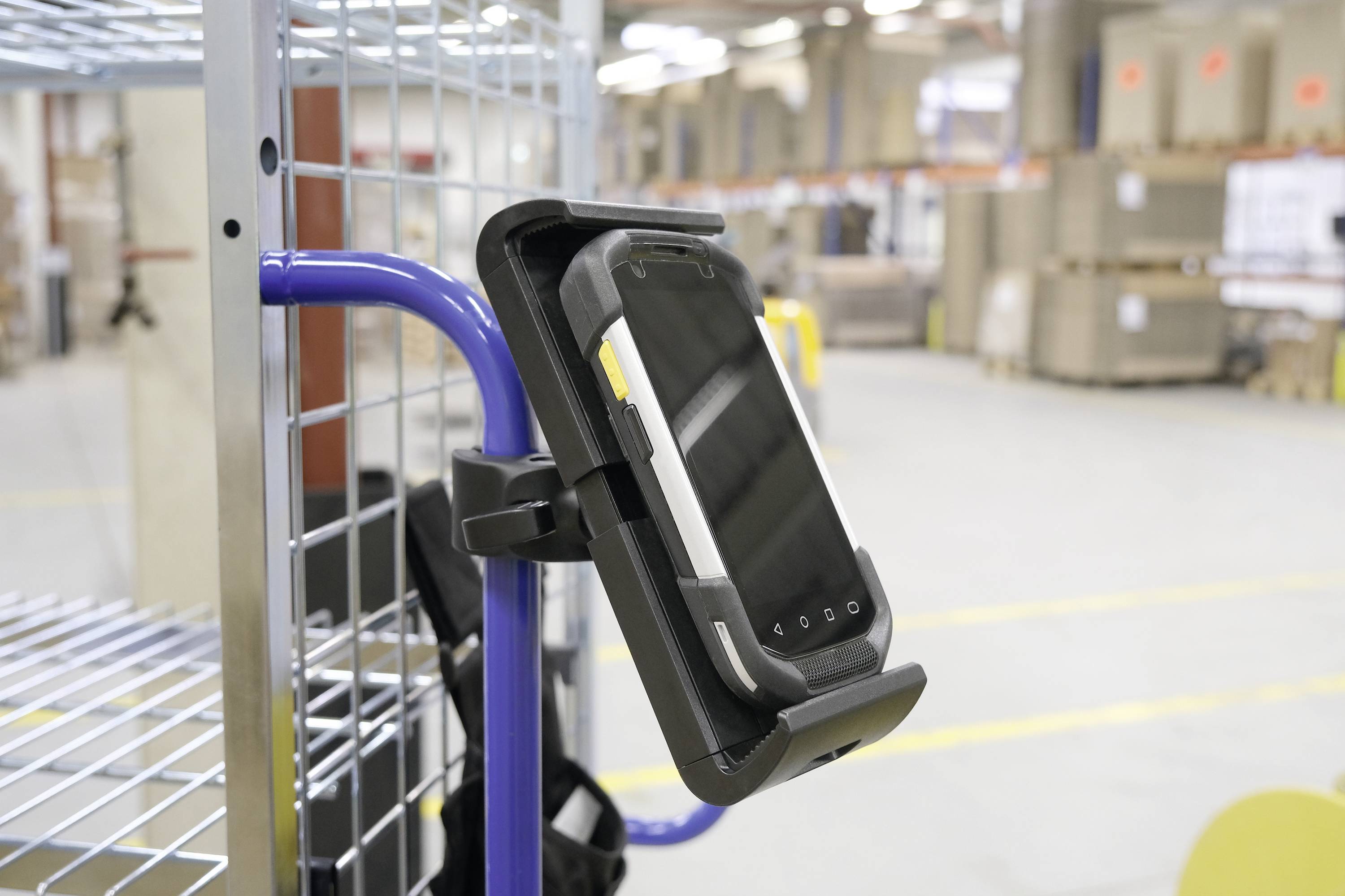 Une tablette dans un support, fixée à un chariot rembourré à l'intérieur d'un grand entrepôt, entourée d'étagères et de cartons.