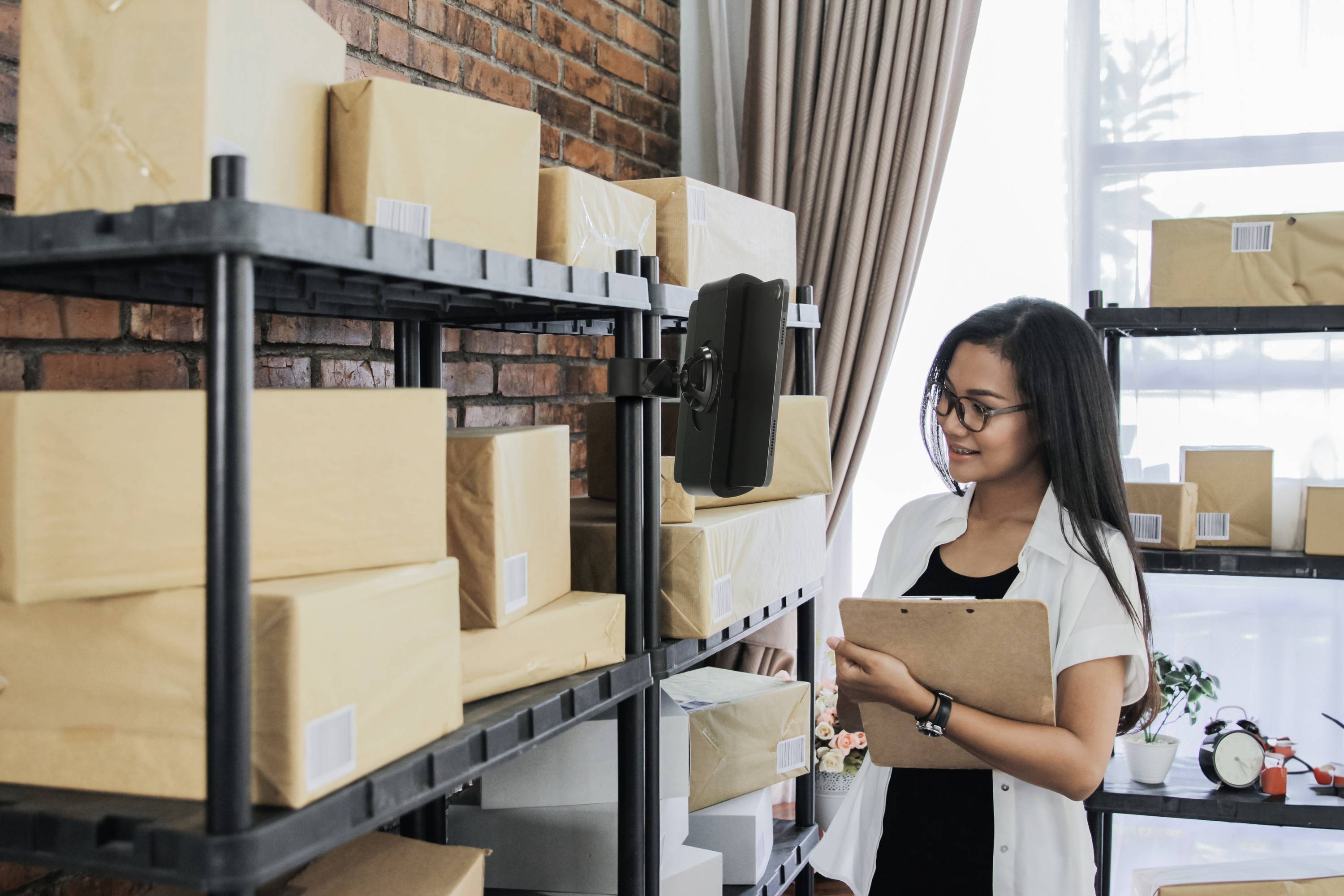 Une personne dans un entrepôt vérifie des notes sur un presse-papiers, entourée de colis empilés sur des étagères métalliques.