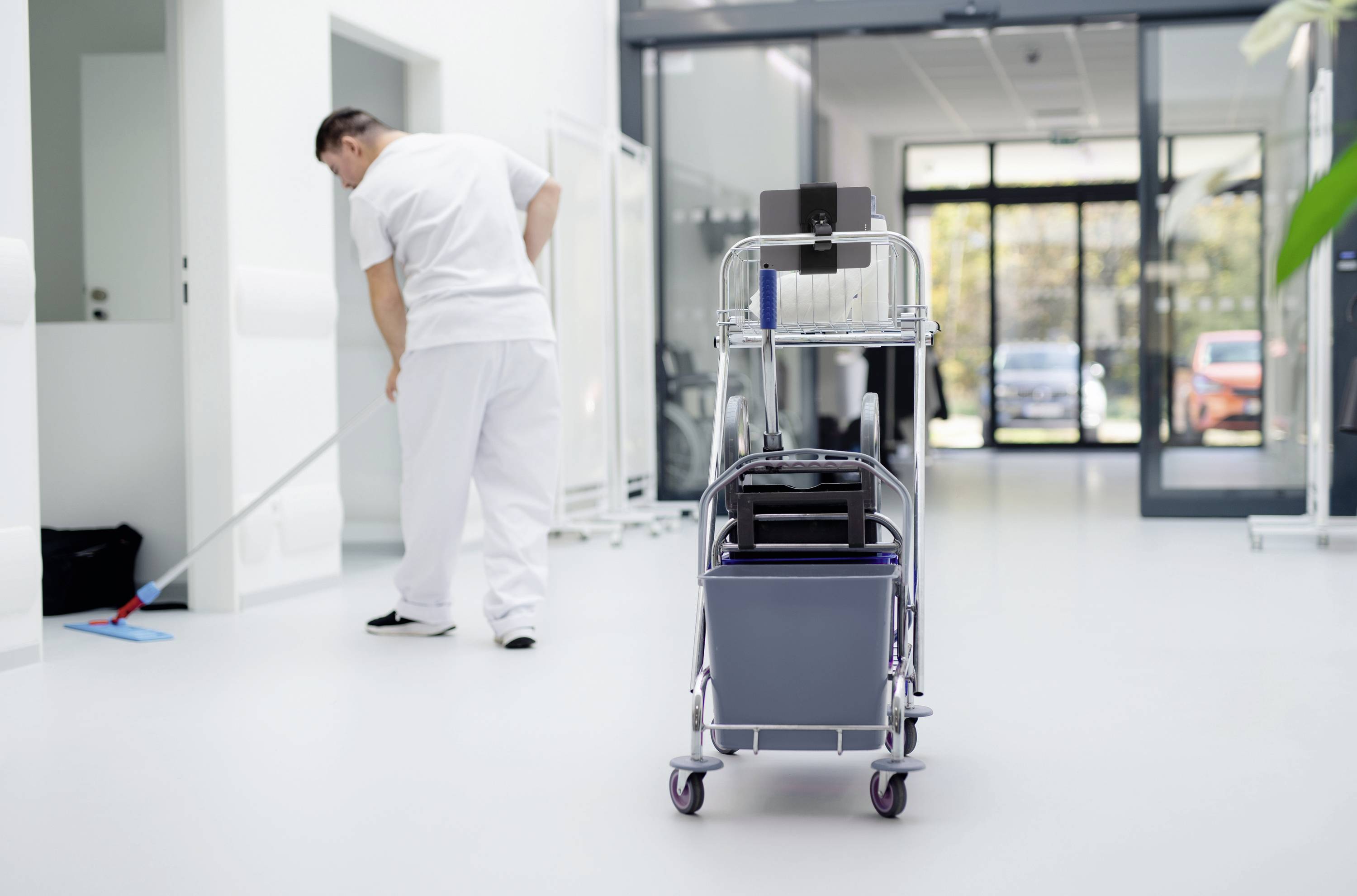 Un agent de nettoyage essuie le sol d'un couloir de bureau moderne et lumineux. Au premier plan, un chariot de nettoyage est équipé de divers ustensiles de nettoyage.