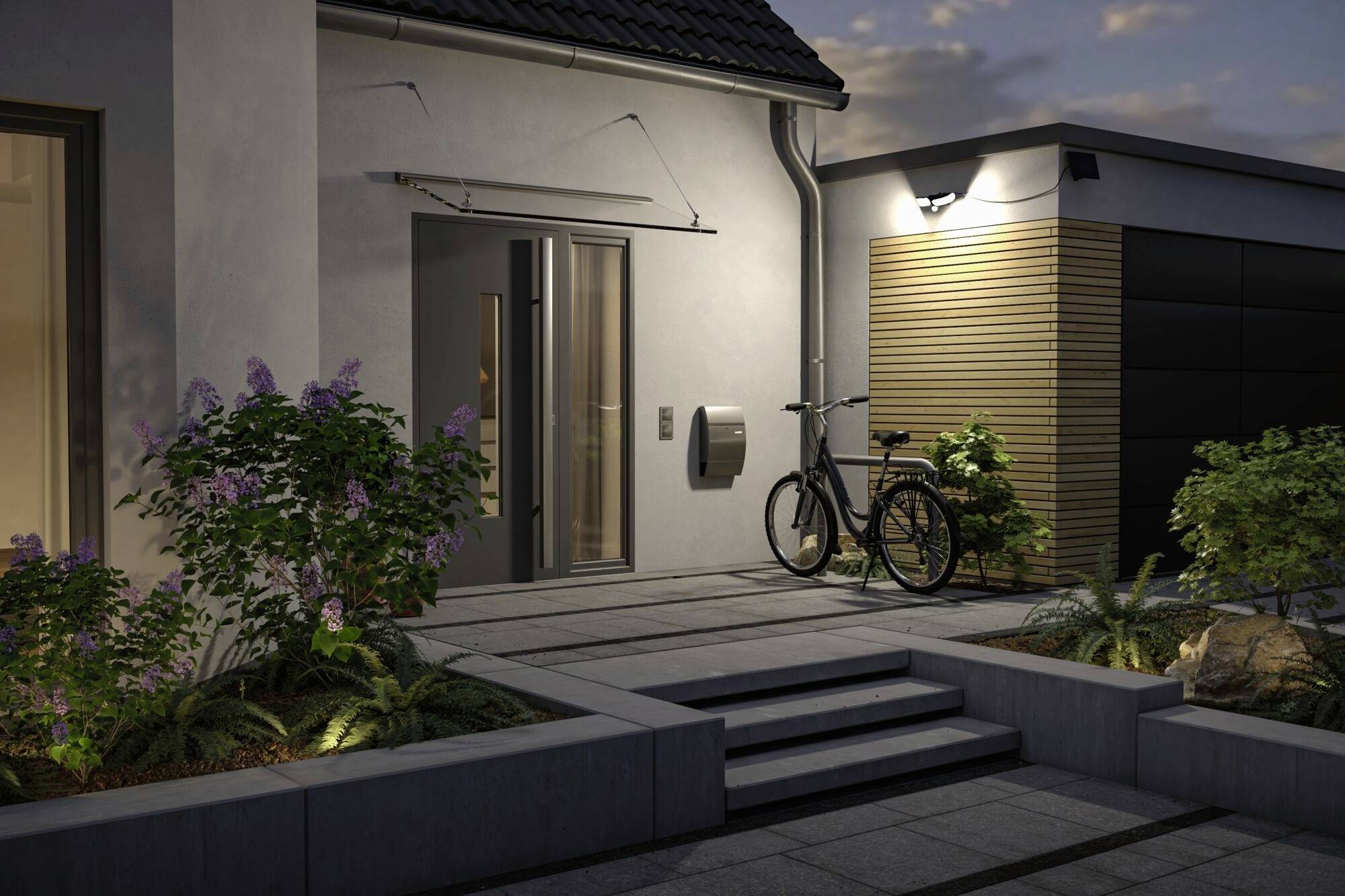 Entrée de maison éclairée au crépuscule avec un vélo à droite de la porte, entourée de parterres de fleurs et d'un chemin pavé.