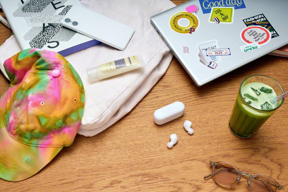 Une casquette colorée, des lunettes de soleil, des écouteurs et une boisson matcha sont posés sur une table à côté d'un ordinateur portable recouvert d'autocollants.