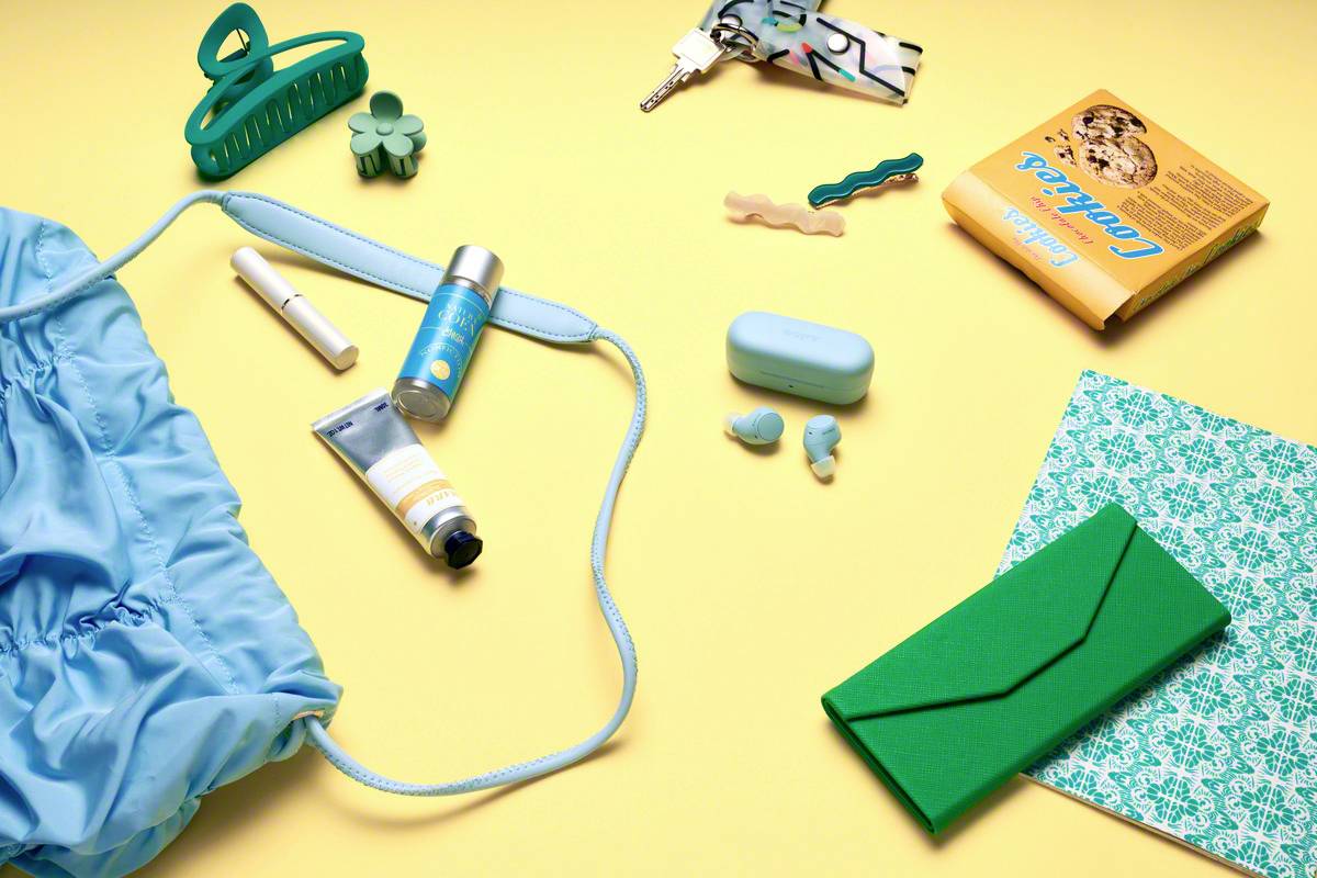 Sur un fond jaune sont dispersés divers objets : un sac bleu, des produits de soin, des clés, un carnet de notes et une trousse verte.