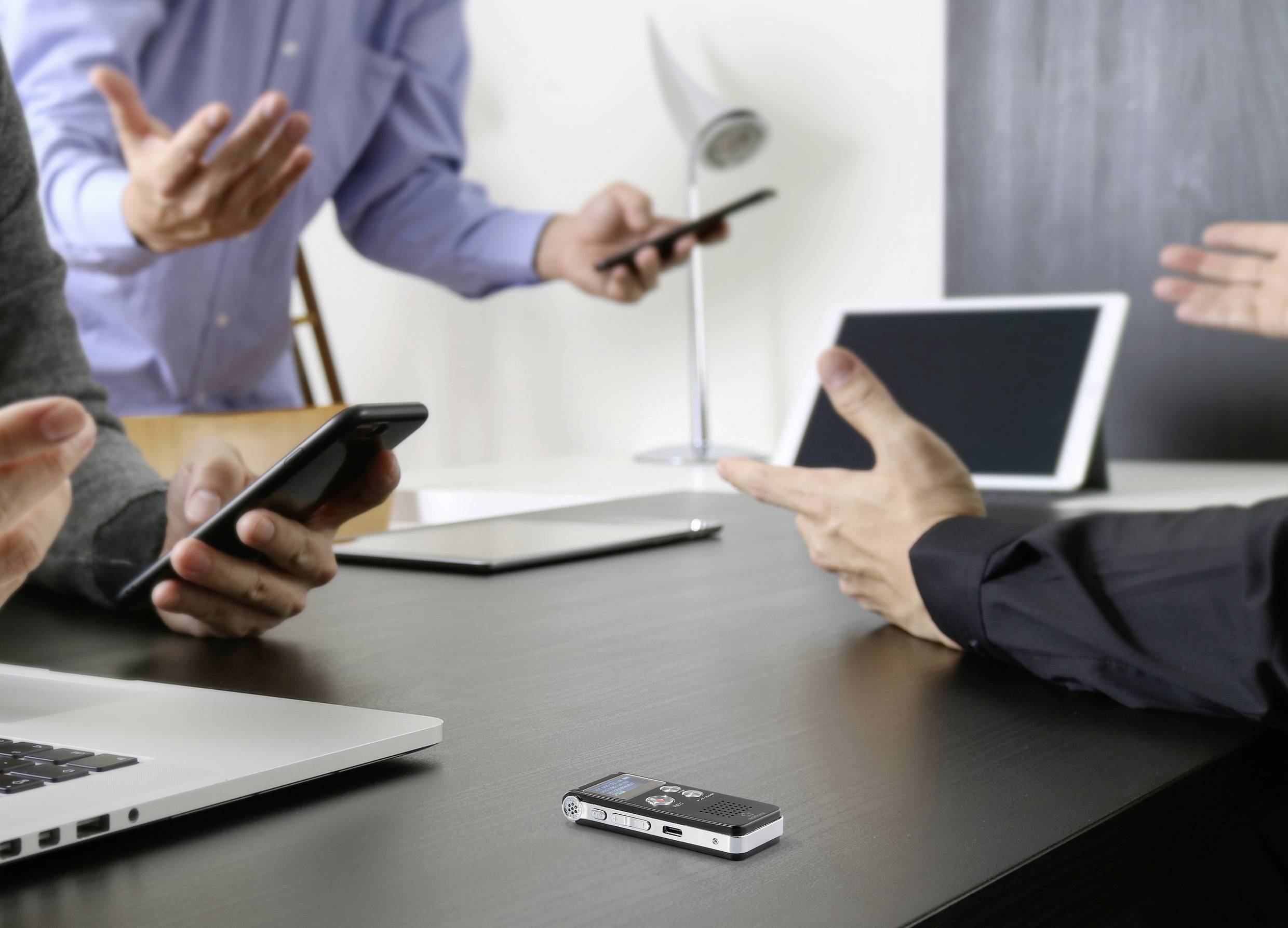 Des personnes discutent dans un bureau, utilisent des ordinateurs portables et des smartphones. Un dictaphone est posé sur la table. L'accent est mis sur la communication digitale.