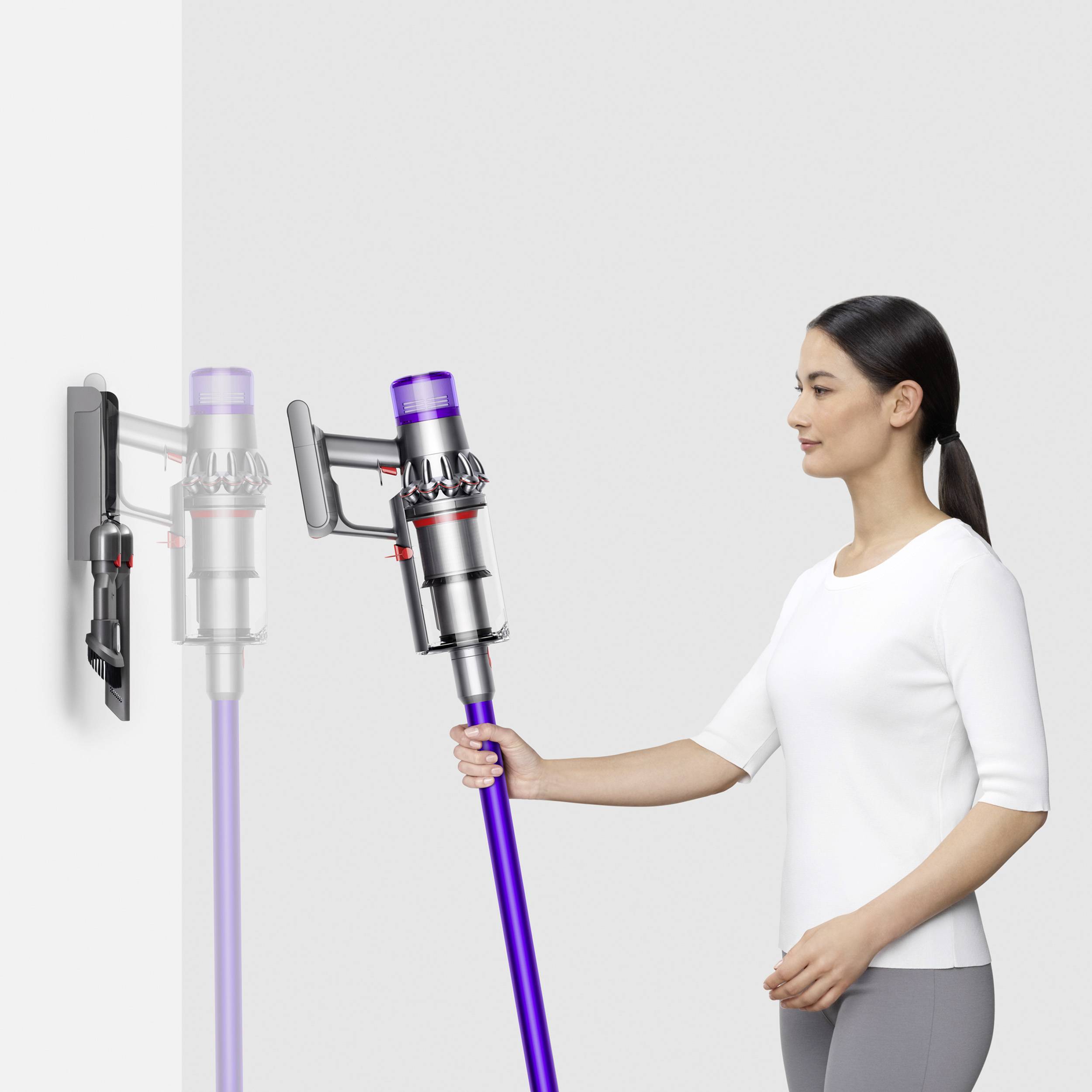 Une personne utilise un aspirateur sans fil, qu'elle prend d'un support mural. L'aspirateur a un tube de couleur lilas.