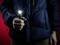 Une personne portant une veste bleue tient une lampe de poche allumée dans sa main devant un fond rouge.