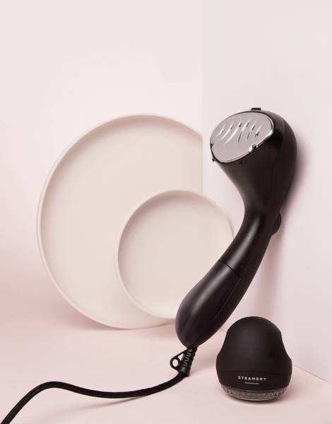 Un nettoyeur vapeur noir à main est appuyé contre un mur rose, à côté de deux assiettes roses et d'un accessoire noir rond.